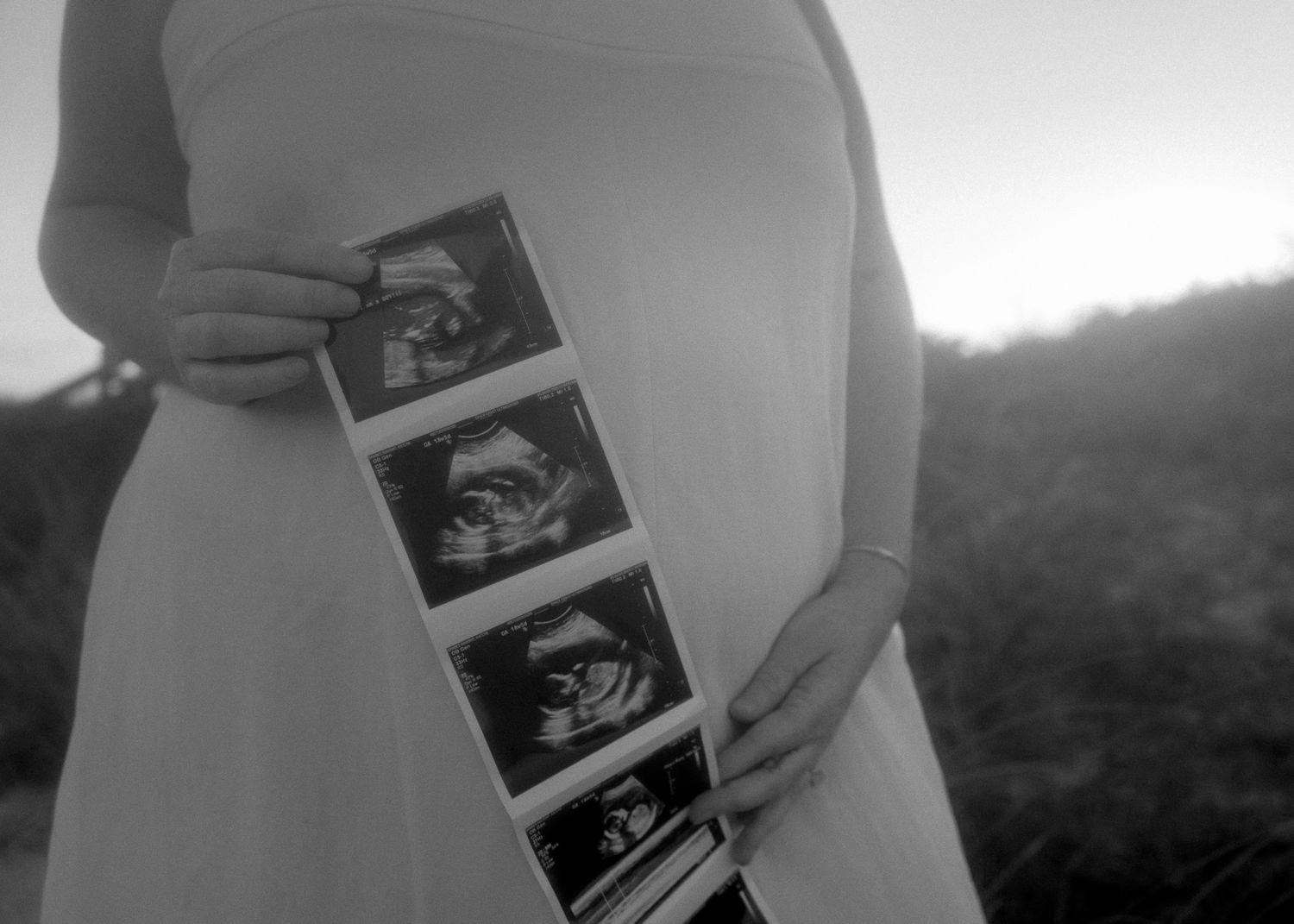 Pregnant woman in white dress holding ultrasound photos outdoors in black and white maternity portrait.