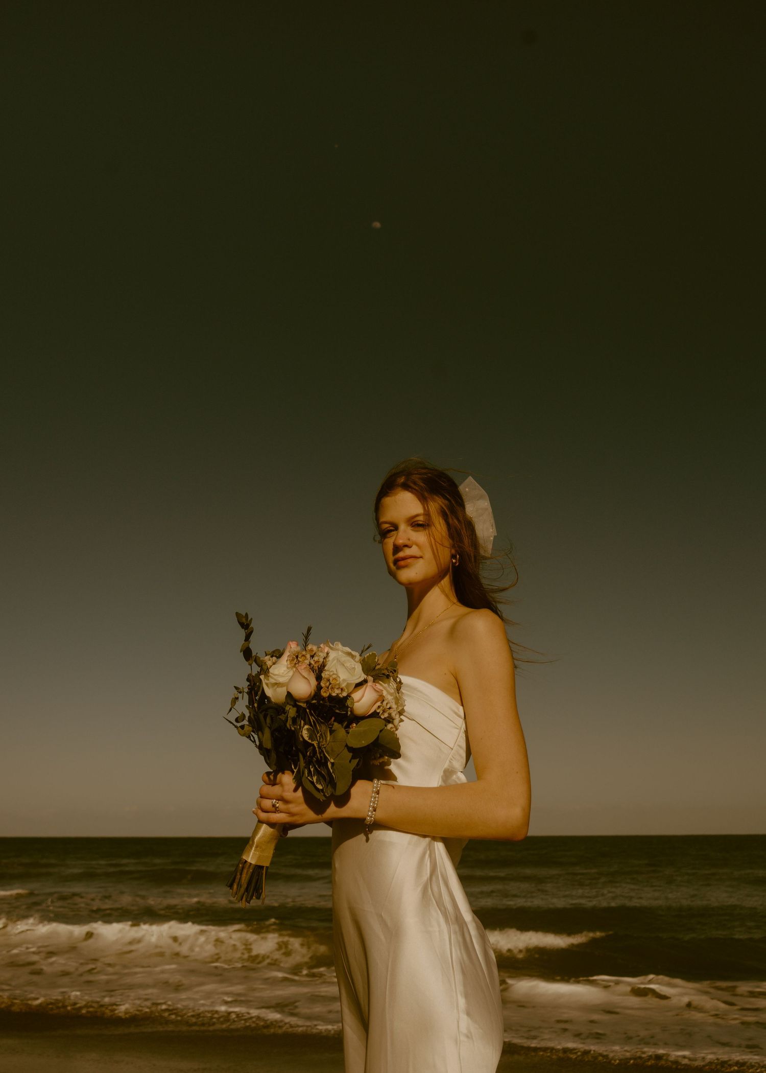 Bride in white gown holding floral bouquet on beach under dramatic evening sky.