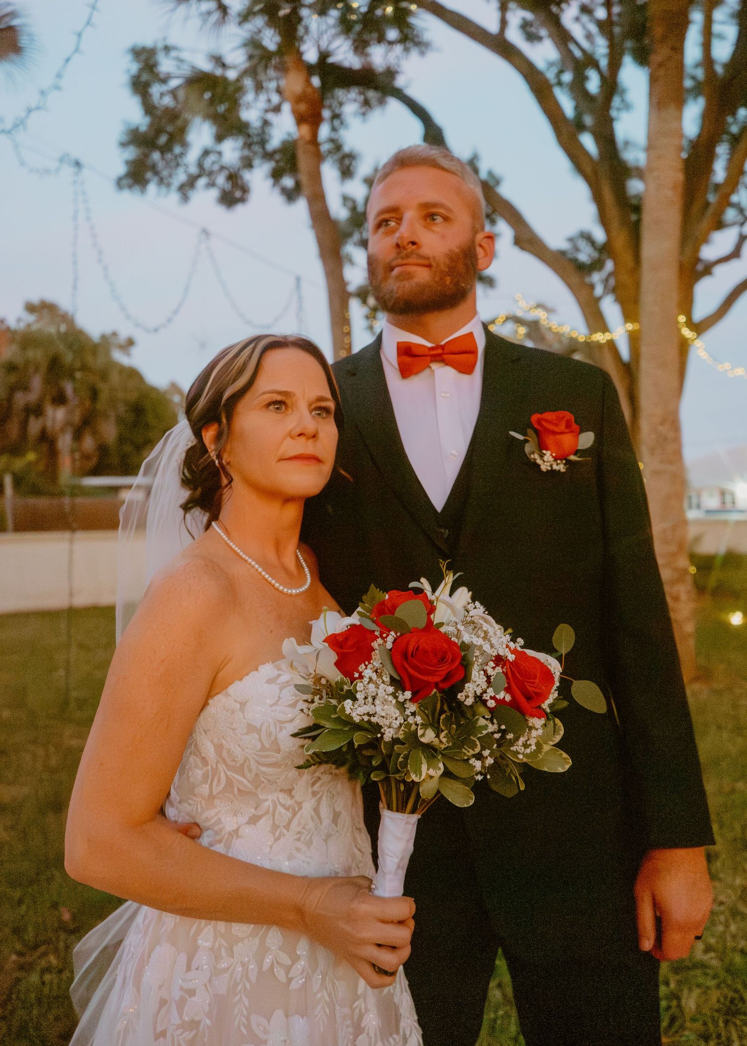 Bride in lace gown holding red rose bouquet stands with groom in black tuxedo and red bow tie outdoors at dusk.