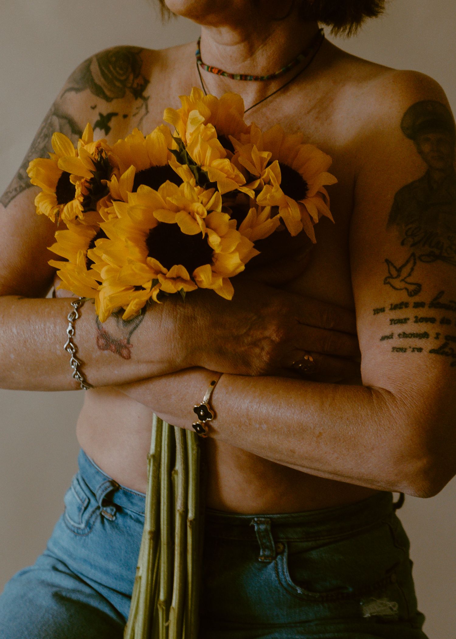 Tattooed person holding vibrant yellow sunflowers against their chest, wearing blue jeans and layered necklaces.