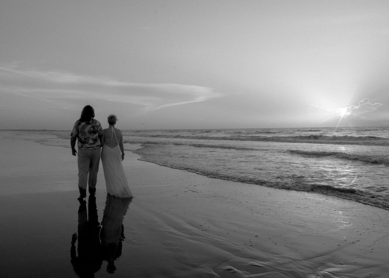 Black and white photo of a couple holding hands on a beach at sunset, waves gently washing ashore.