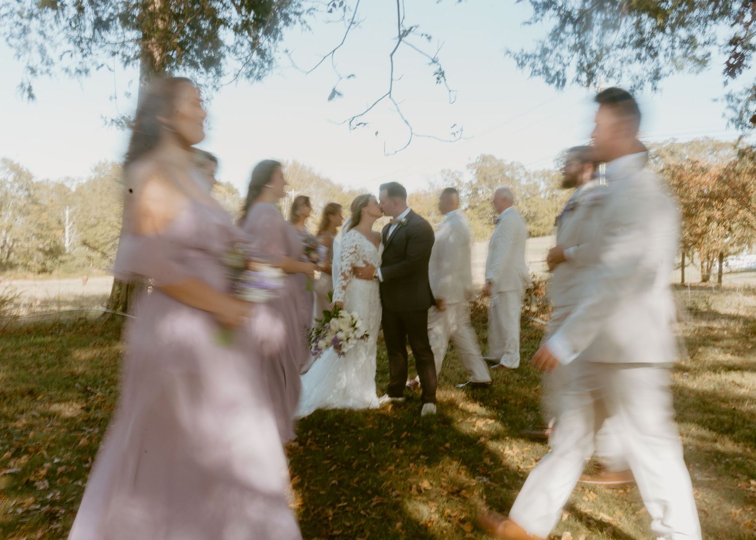 Blurred wedding party in lavender and white attire gathered outdoors under trees during a sunlit ceremony.
