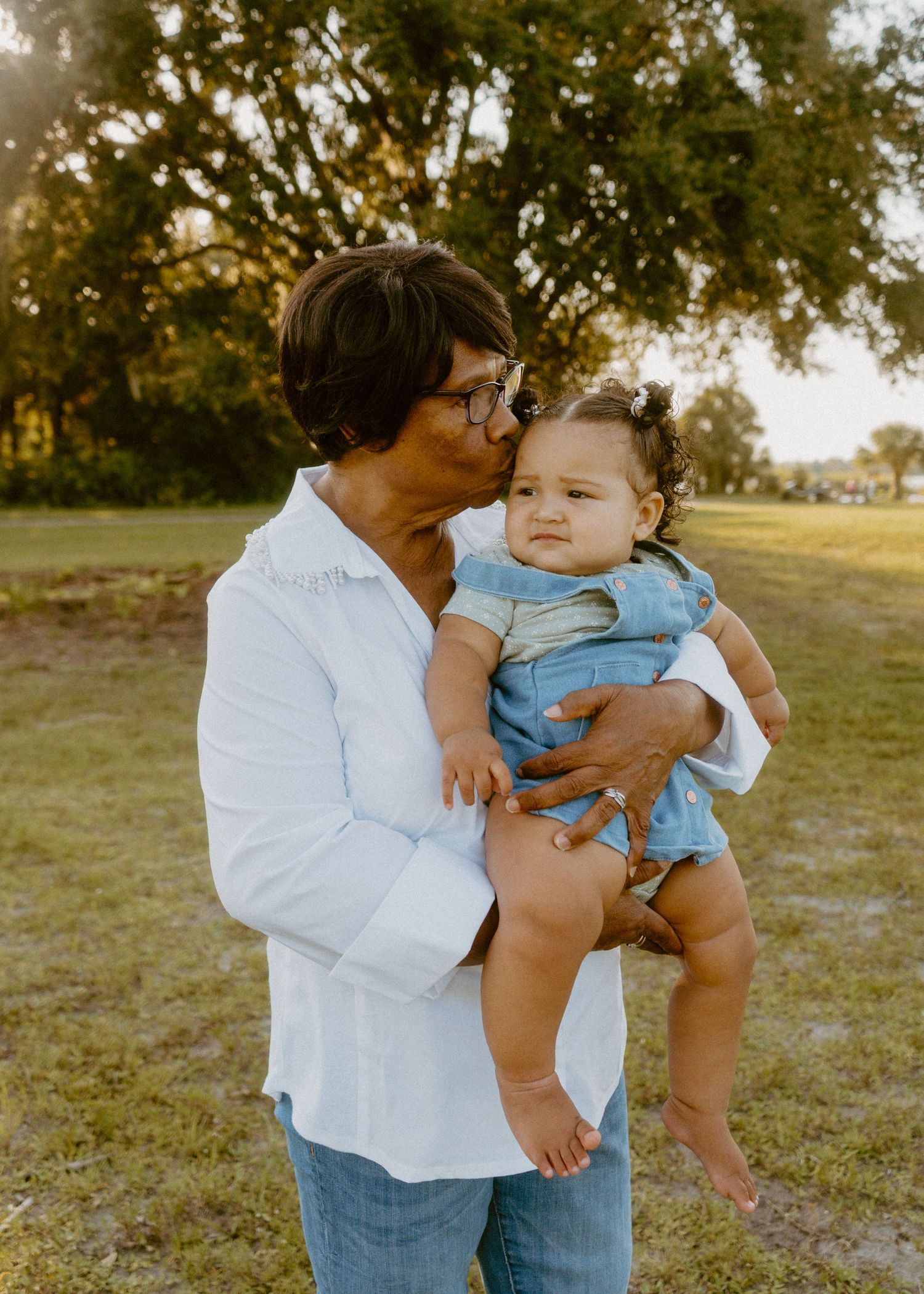 Adult kissing a baby in denim overalls outdoors in a sunny park setting.