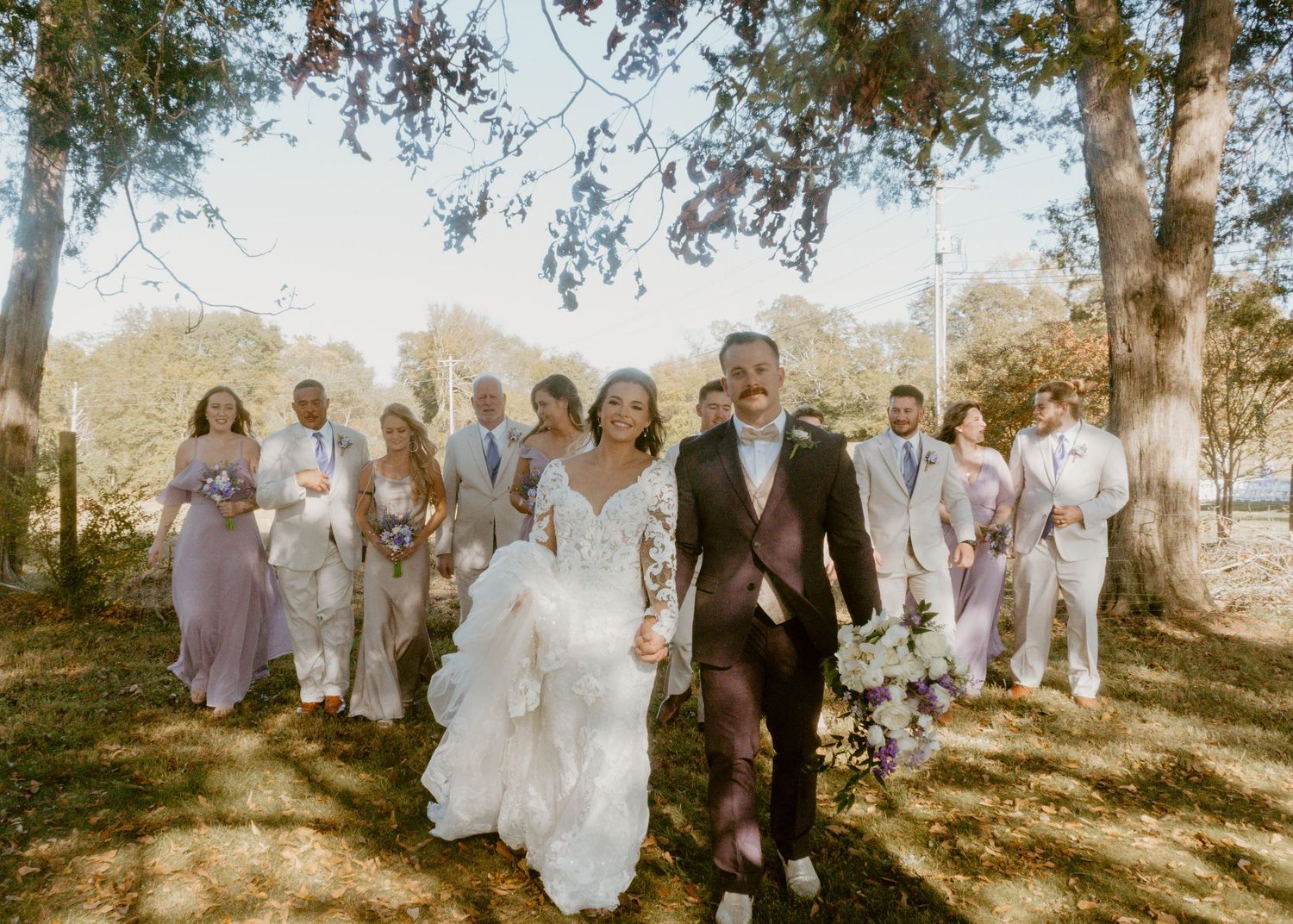 Bride and groom lead wedding party through tree-lined outdoor venue on sunny day.