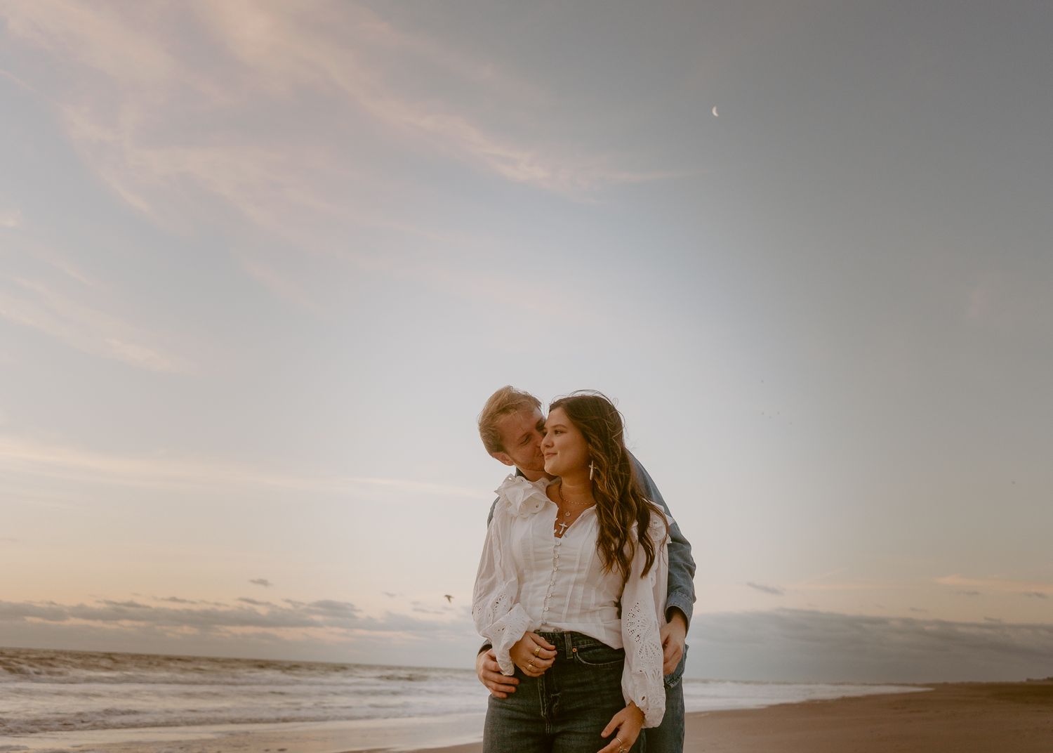 Couple embracing romantically on a sandy beach at golden hour with ocean waves and pastel sky behind them.