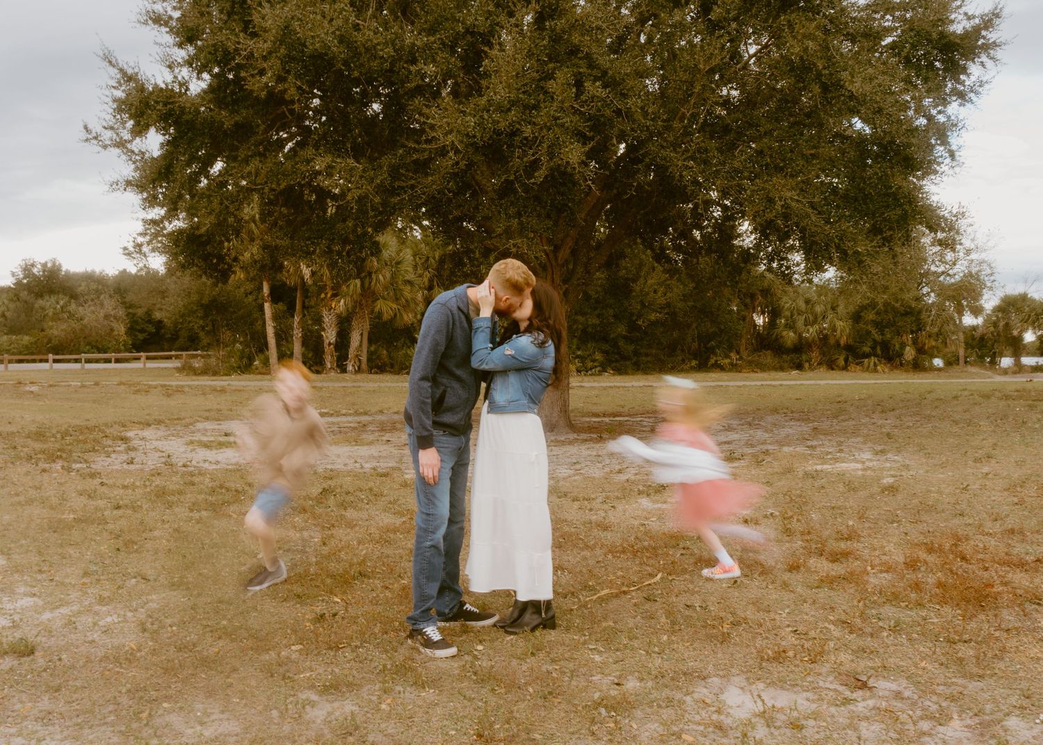Couple embracing in a field with children running around them, blurred motion, large trees in background.