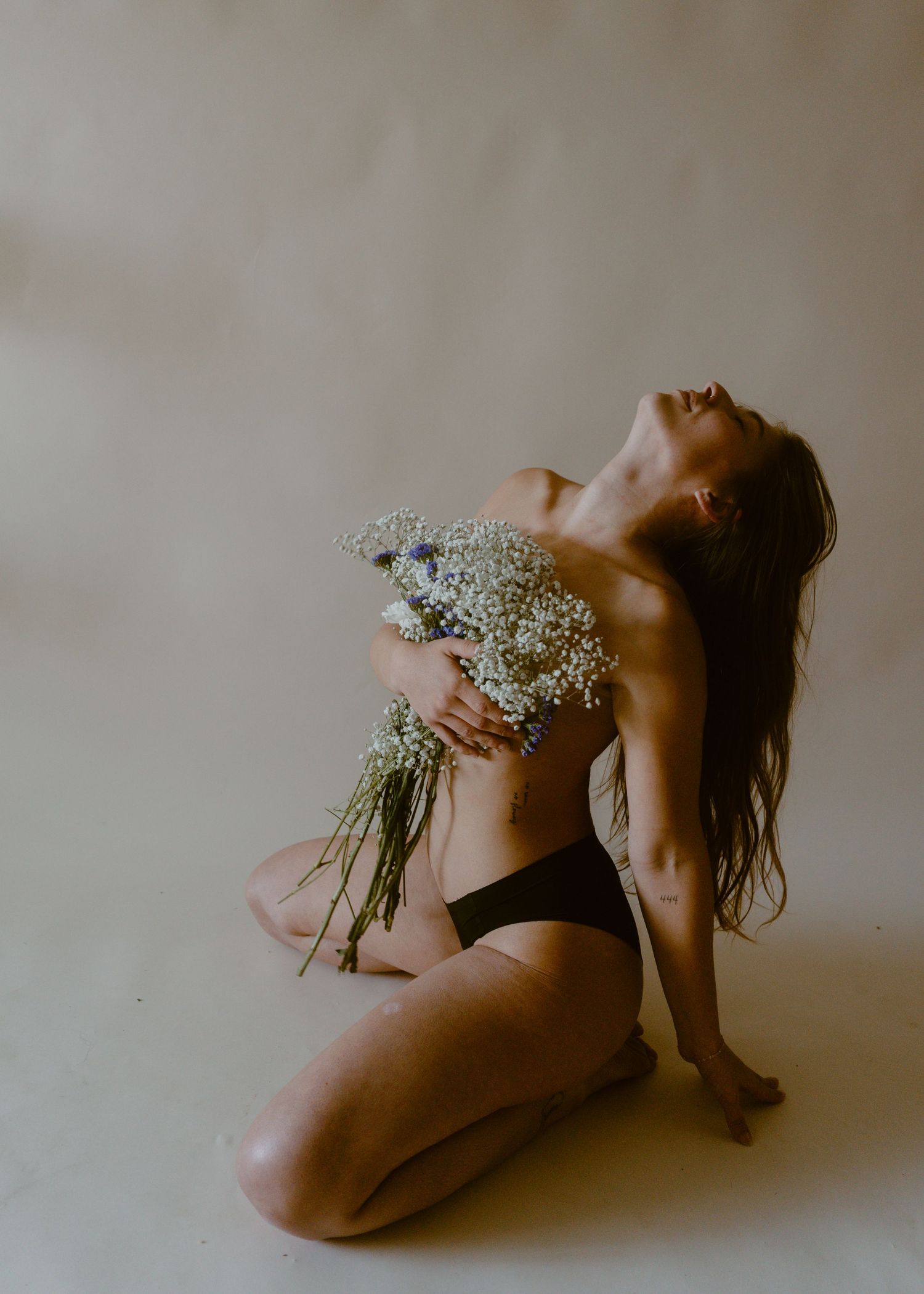 Woman kneeling on studio floor holding white wildflowers, wearing black underwear, head tilted back with flowing hair.