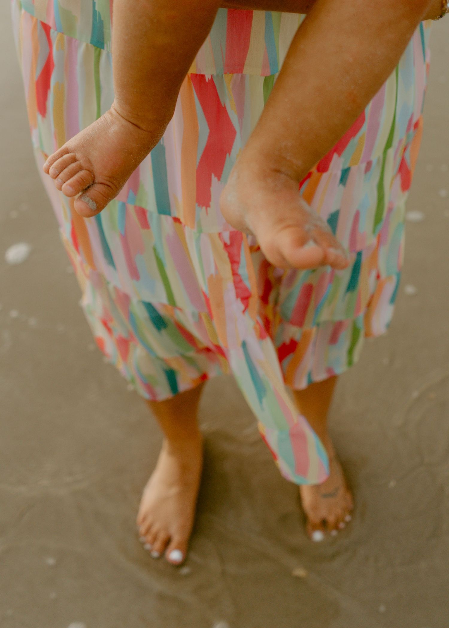 Adult and baby bare feet standing on wet sand, wearing a colorful brushstroke-print tiered skirt at the beach.