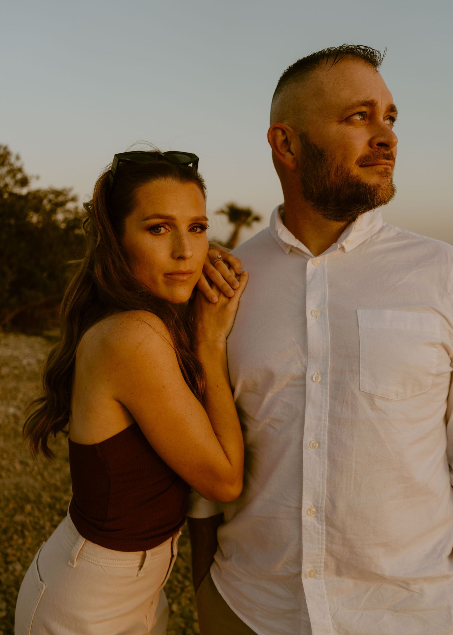 A couple poses outdoors at golden hour, woman in brown top with sunglasses, man in white shirt gazing away.