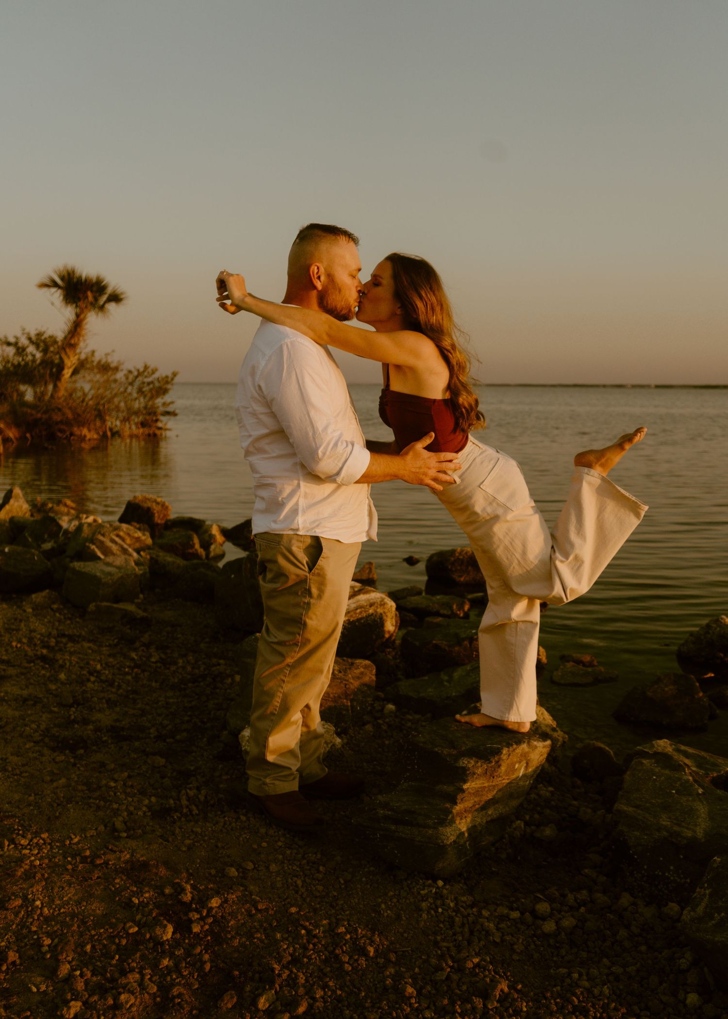 Couple sharing a romantic kiss at golden hour on a rocky shoreline with calm water and palm trees in the background.