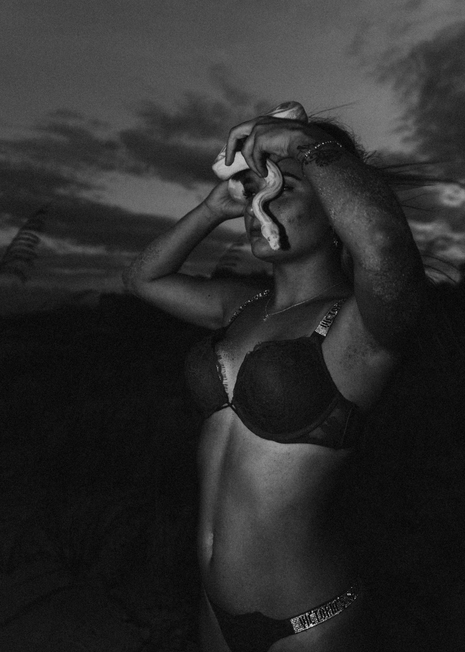 Black and white photo of a woman in bikini holding a snake against dramatic sunset sky.