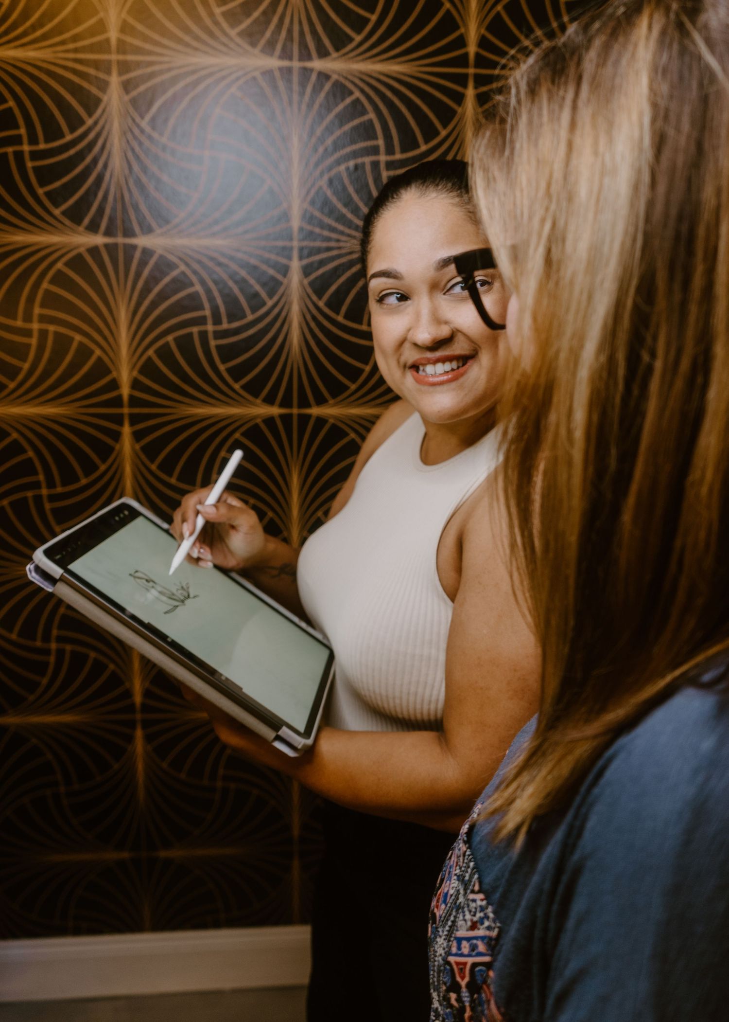 A smiling woman holds an iPad with an Apple Pencil, showing content to another person against a patterned wall.