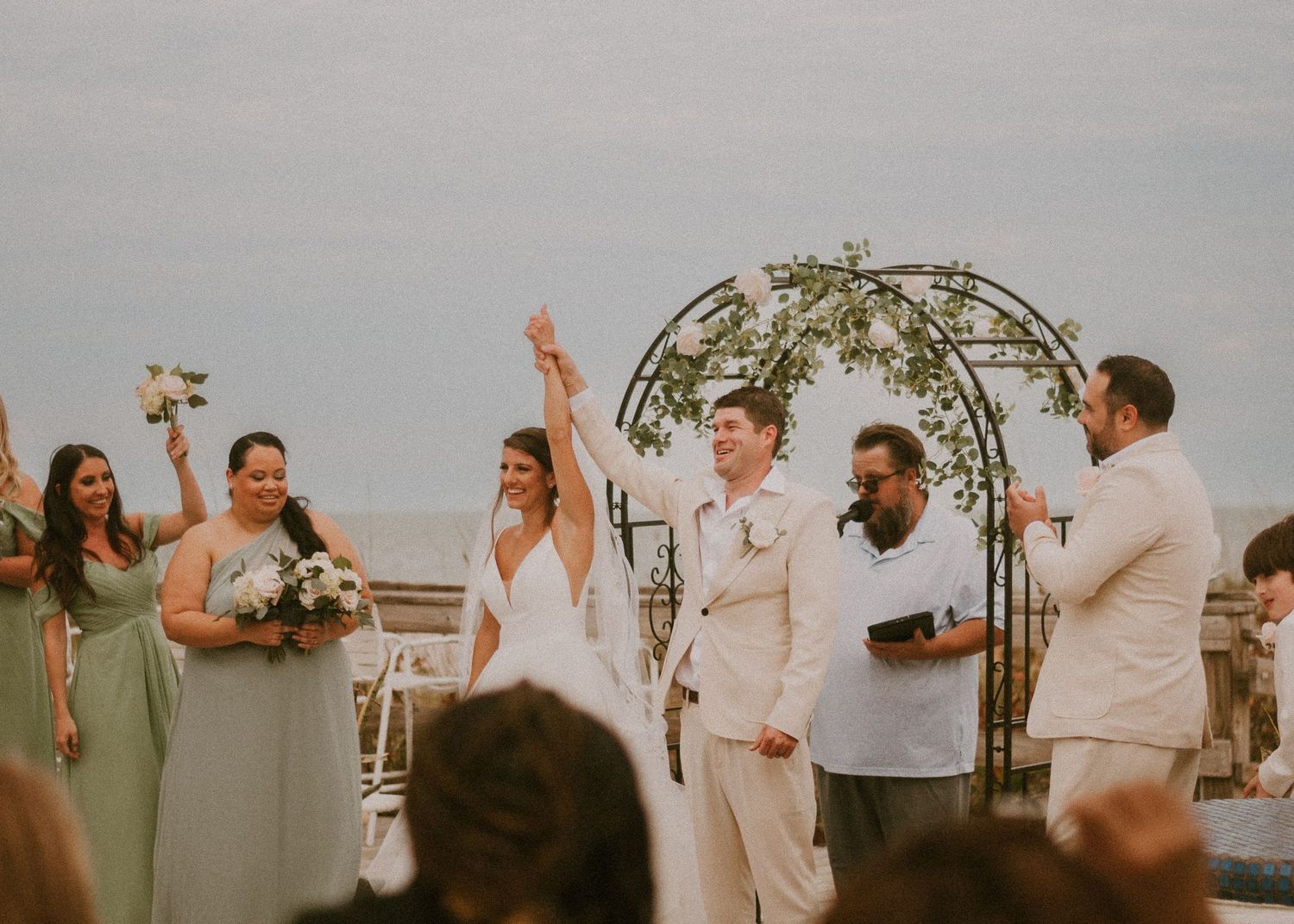 Joyful bride raising arm at outdoor beach wedding ceremony with bridal party.