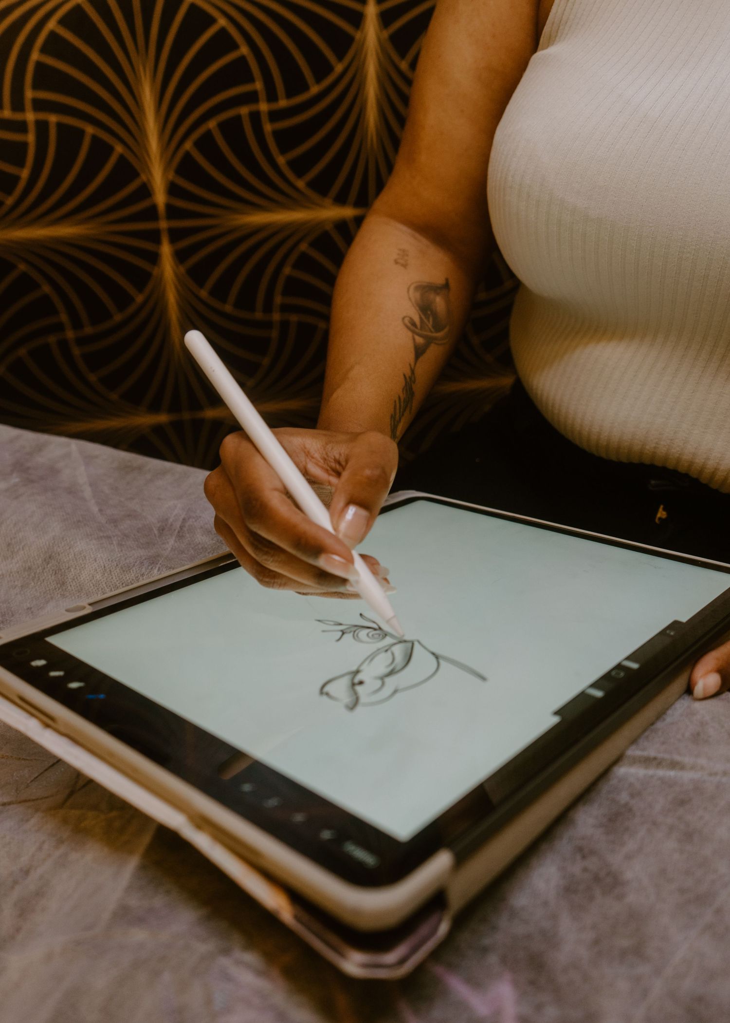A tattooed woman draws botanical art on a tablet using a stylus, wearing a white ribbed top.