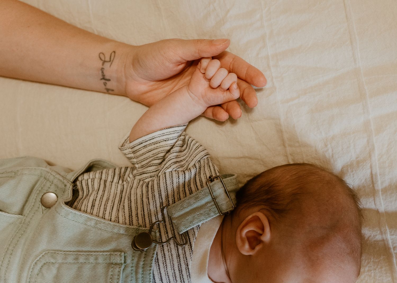 Newborn baby holding mother's finger, lying on white bedding, wearing striped onesie, close-up bonding moment.