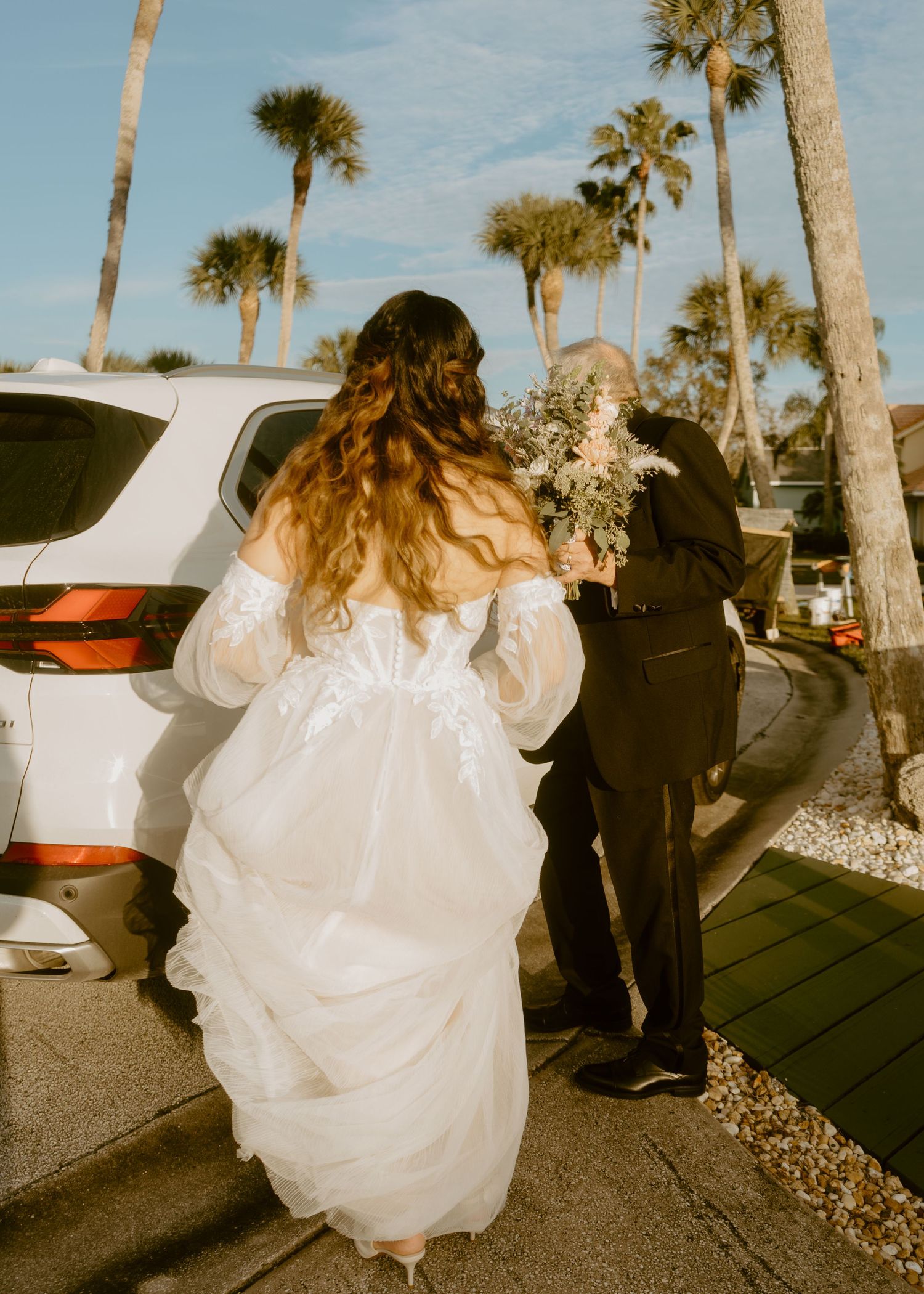 Bride in off-shoulder lace wedding gown walking with groom near palm trees holding bouquet.