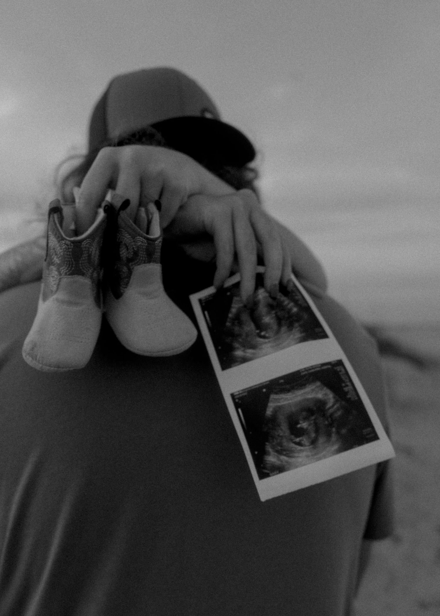 Black and white pregnancy announcement with baby shoes and ultrasound photos at beach.