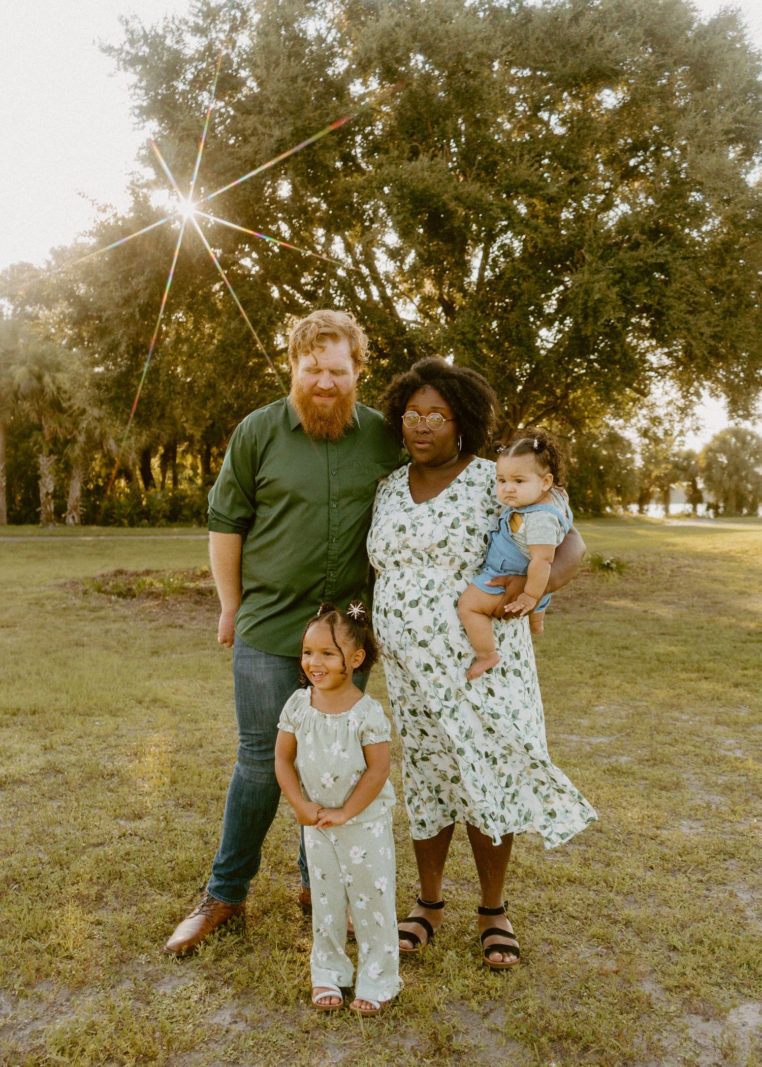 Family of four poses outdoors at golden hour, parents with two young children on green grass with trees behind them.