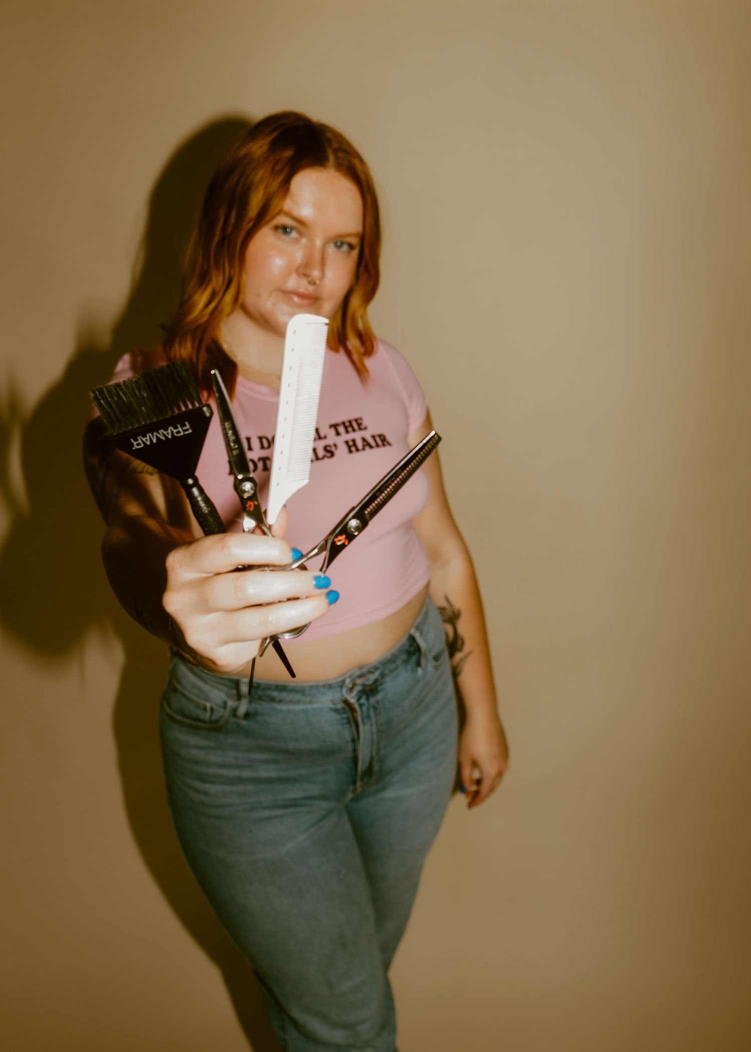 Young woman with red hair holding hair scissors and styling tools, wearing a pink crop top and jeans.