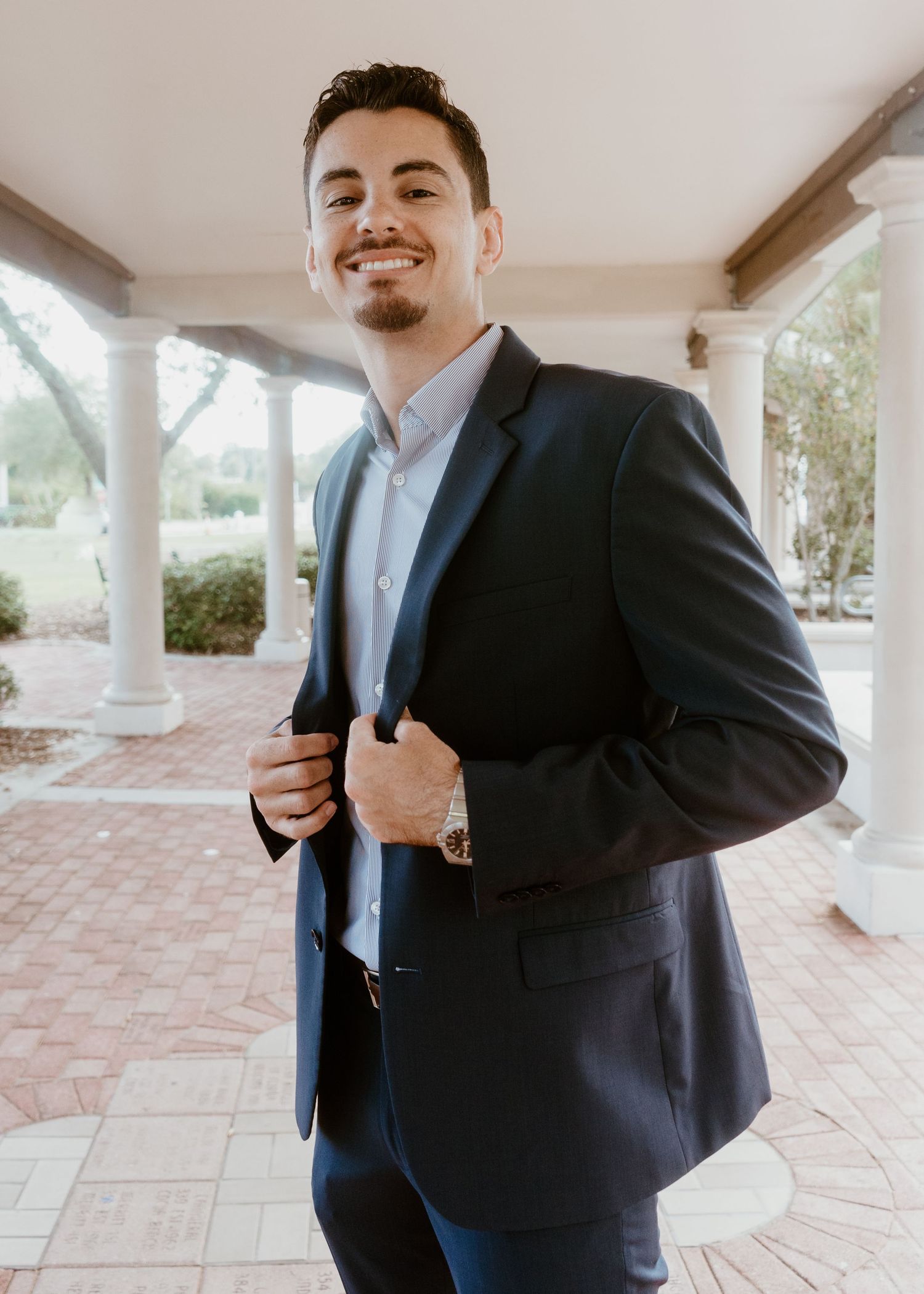 Young man in navy blazer smiling outdoors near white columns on a brick patio.