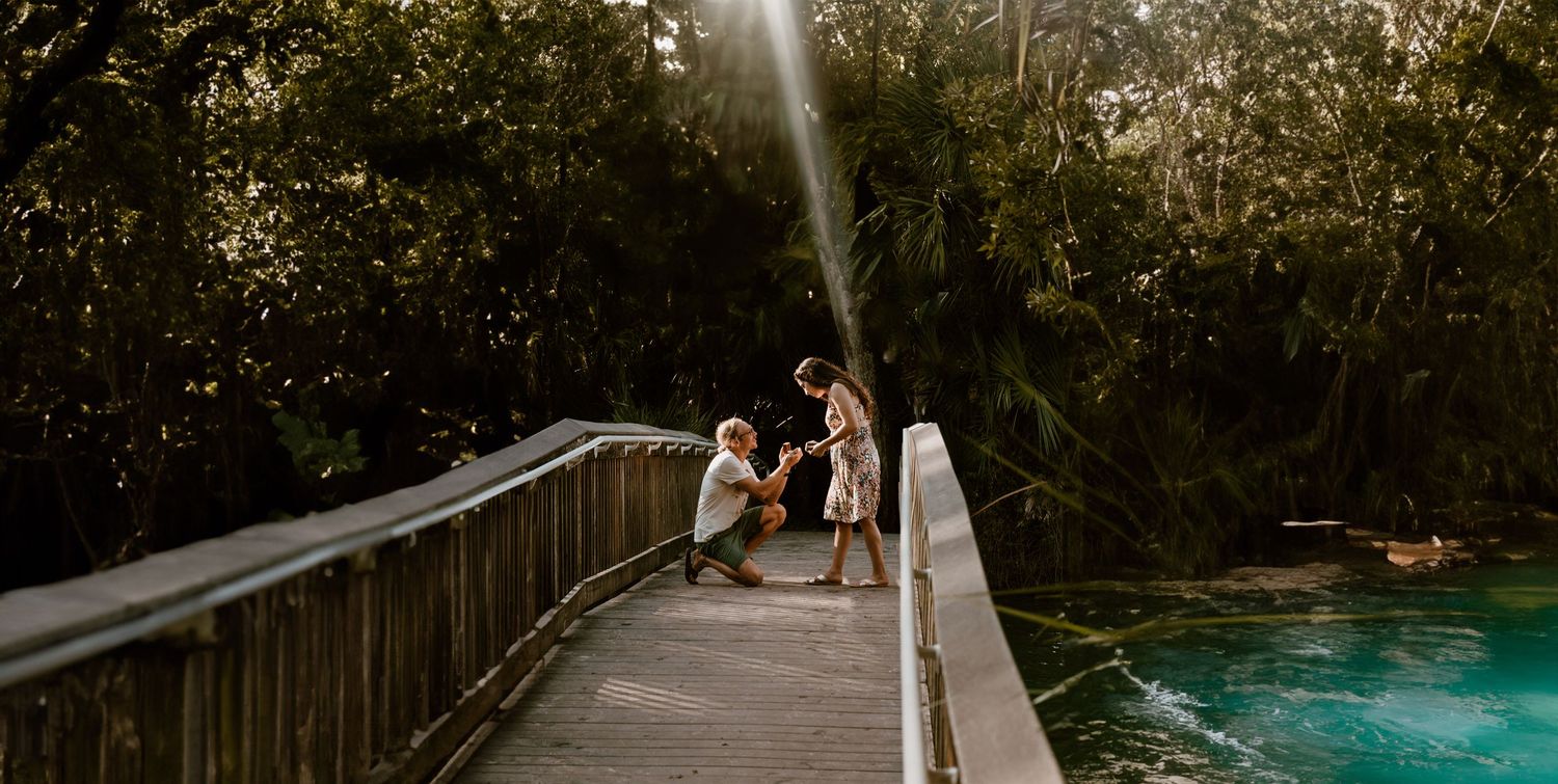 Couple on a wooden bridge surrounded by lush green trees near turquoise water in a tropical setting.