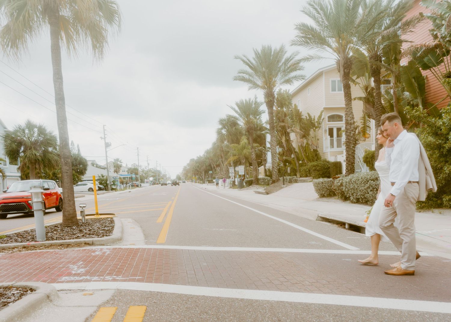 A man in a white shirt crosses a sunny palm-lined street in a tropical coastal town with colorful buildings nearby.