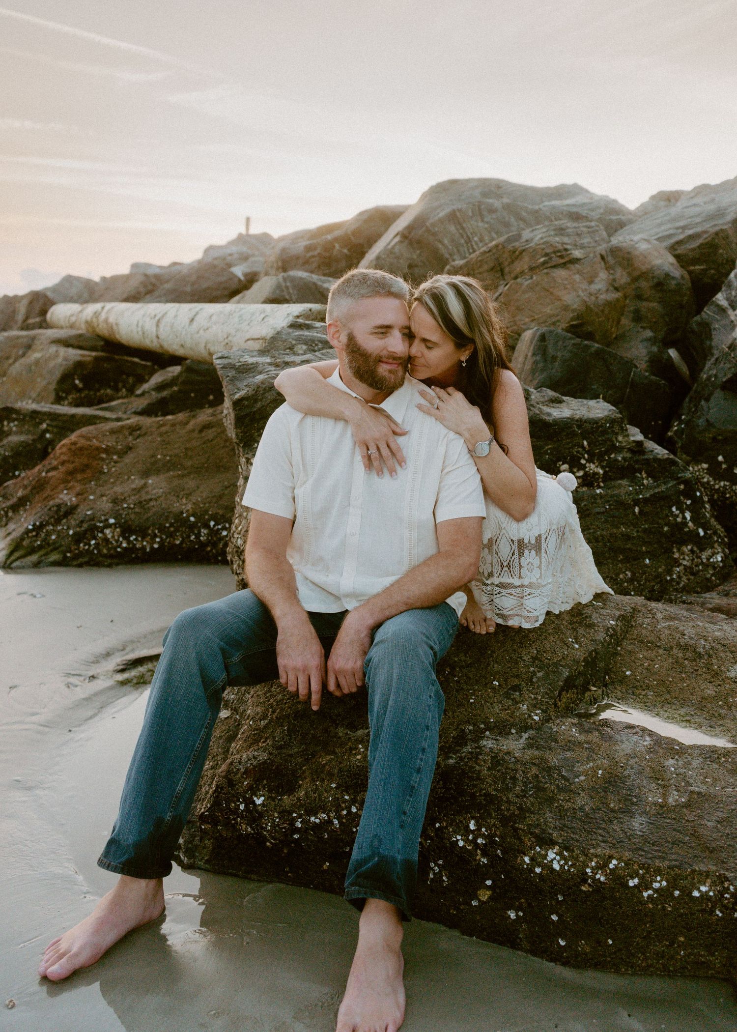 Couple sitting closely together on rocks at the beach during a warm sunset.