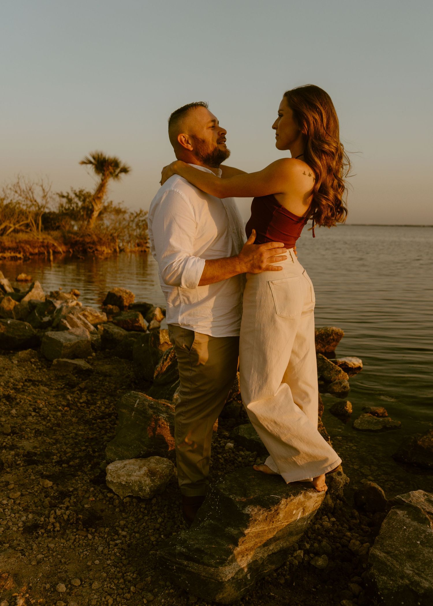 Couple embracing romantically on rocky shoreline at golden hour sunset near calm ocean waters.