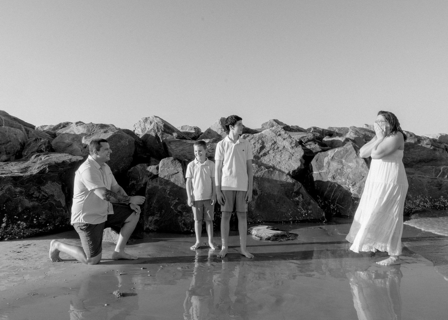 Black and white beach photo of family with man kneeling before woman and two children near rocks.