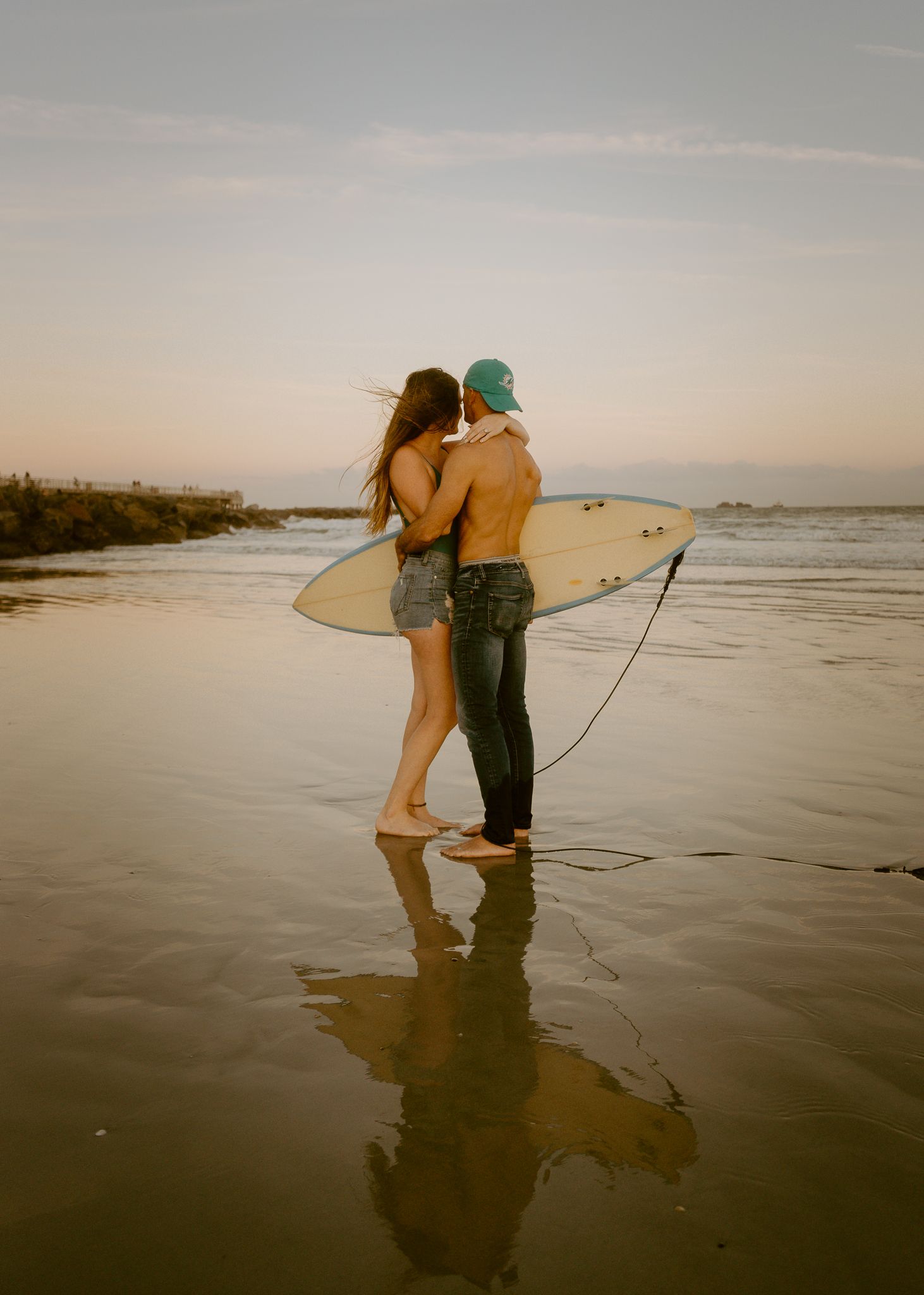 Couple sharing a romantic kiss on the beach at sunset while holding a white surfboard near the ocean.