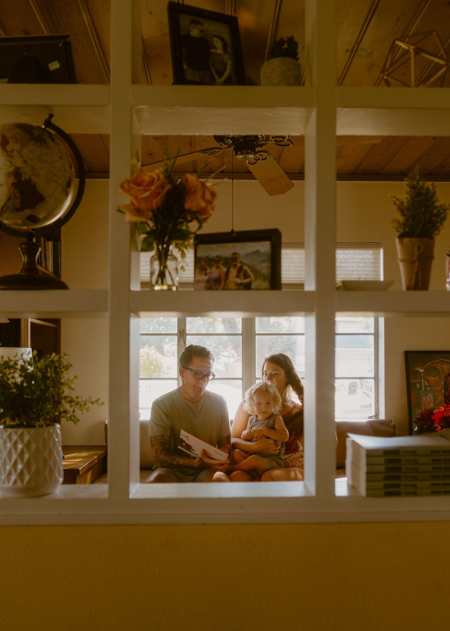 Family reading together on a cozy window seat, surrounded by shelves with globe, flowers, and framed photos in a warm home.