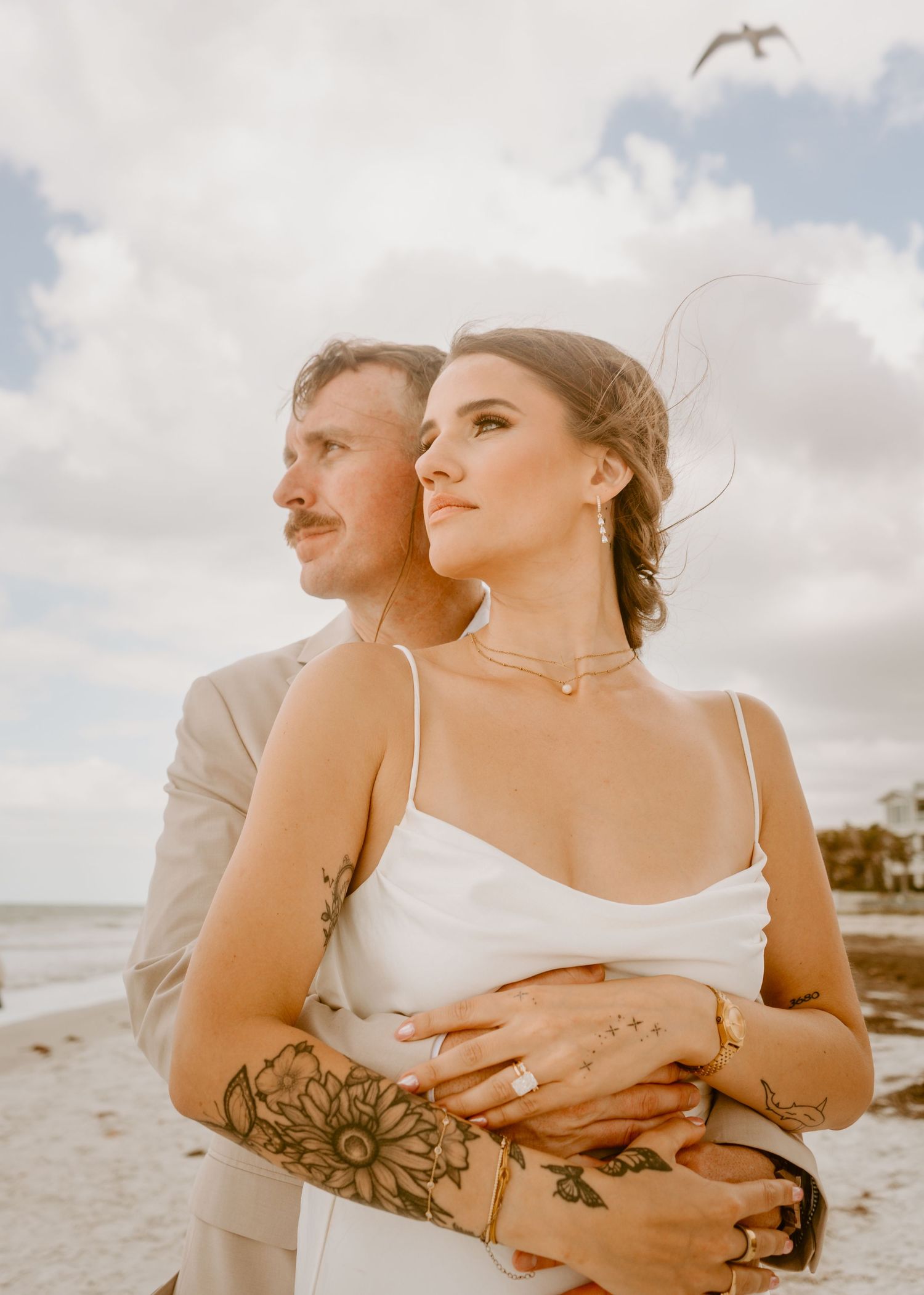 Couple embracing on beach, bride in white dress with floral tattoo, groom in tan suit, cloudy sky backdrop.