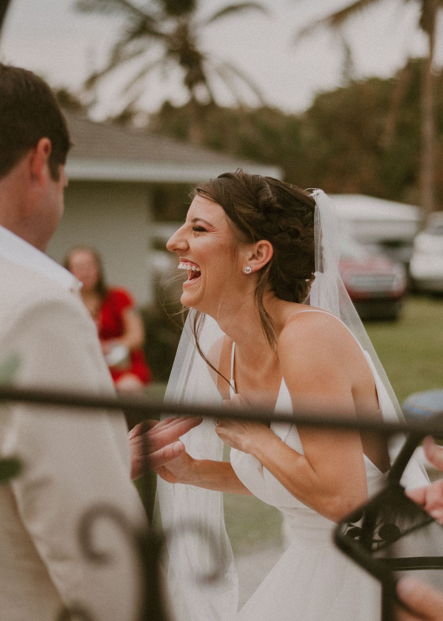 Joyful bride in white gown and veil laughing during outdoor wedding ceremony with groom in tan suit.