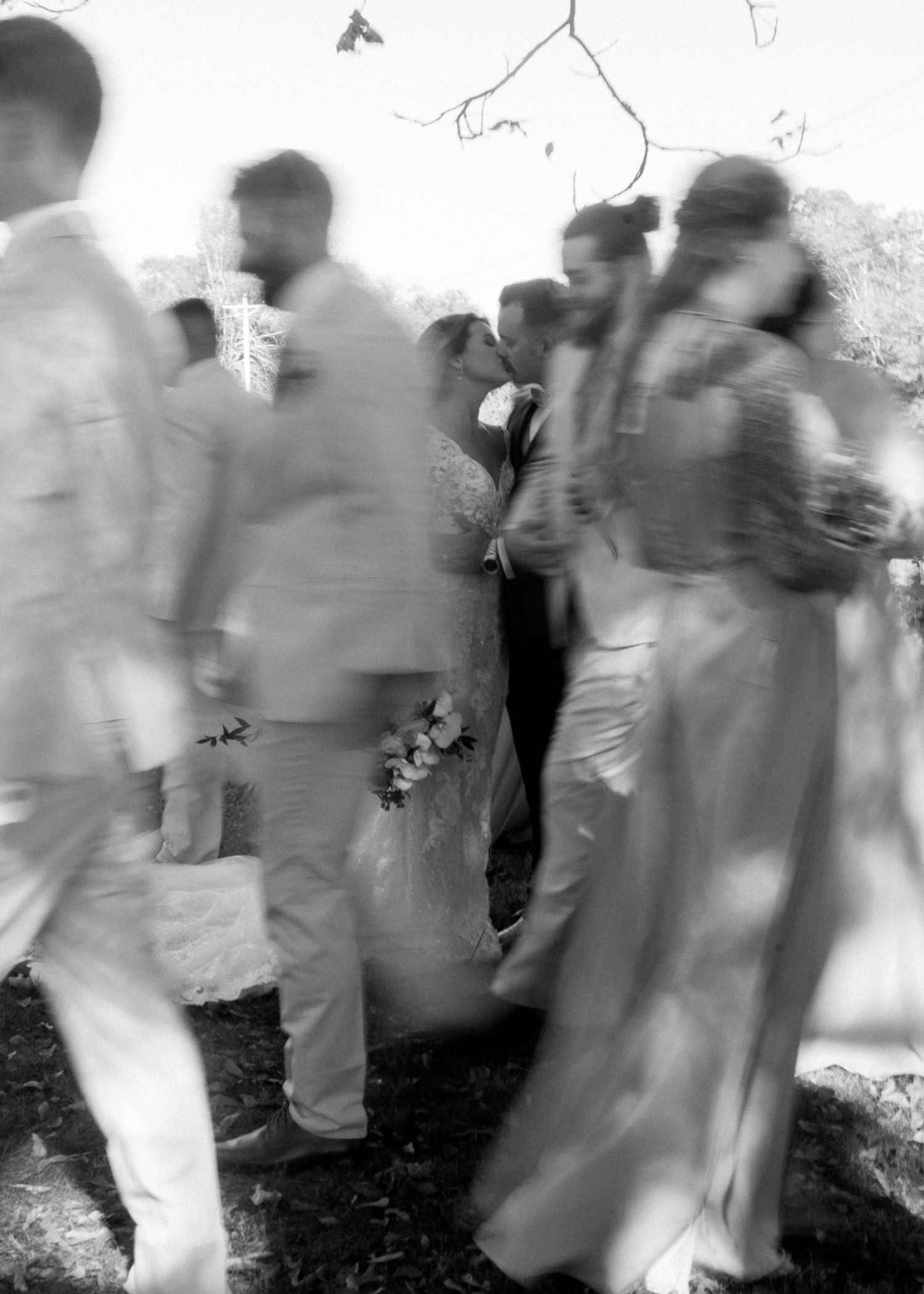 Black and white wedding photo of bride and groom kissing surrounded by guests outdoors.