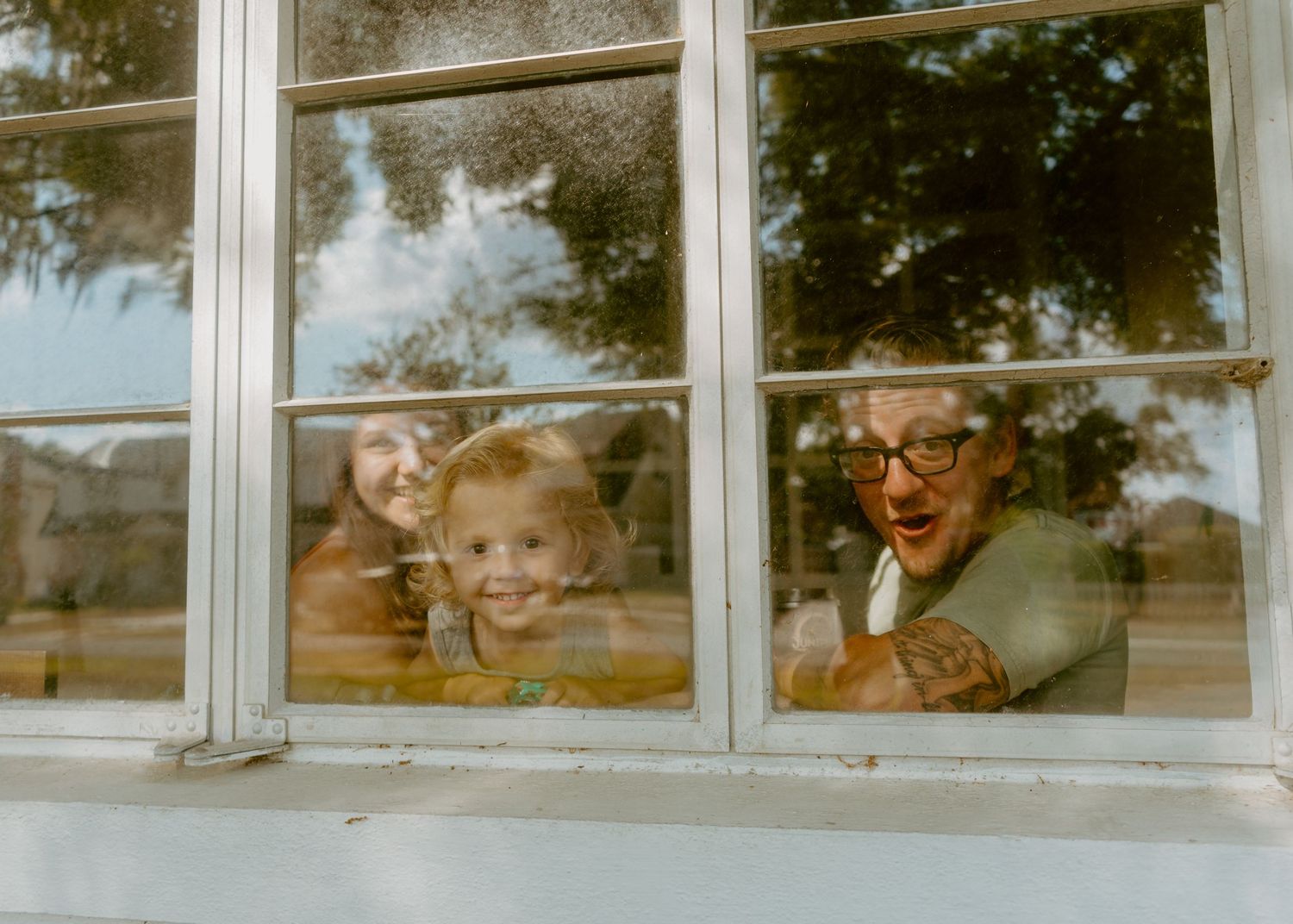 Family of three smiling and waving through a white-framed window surrounded by lush green trees.