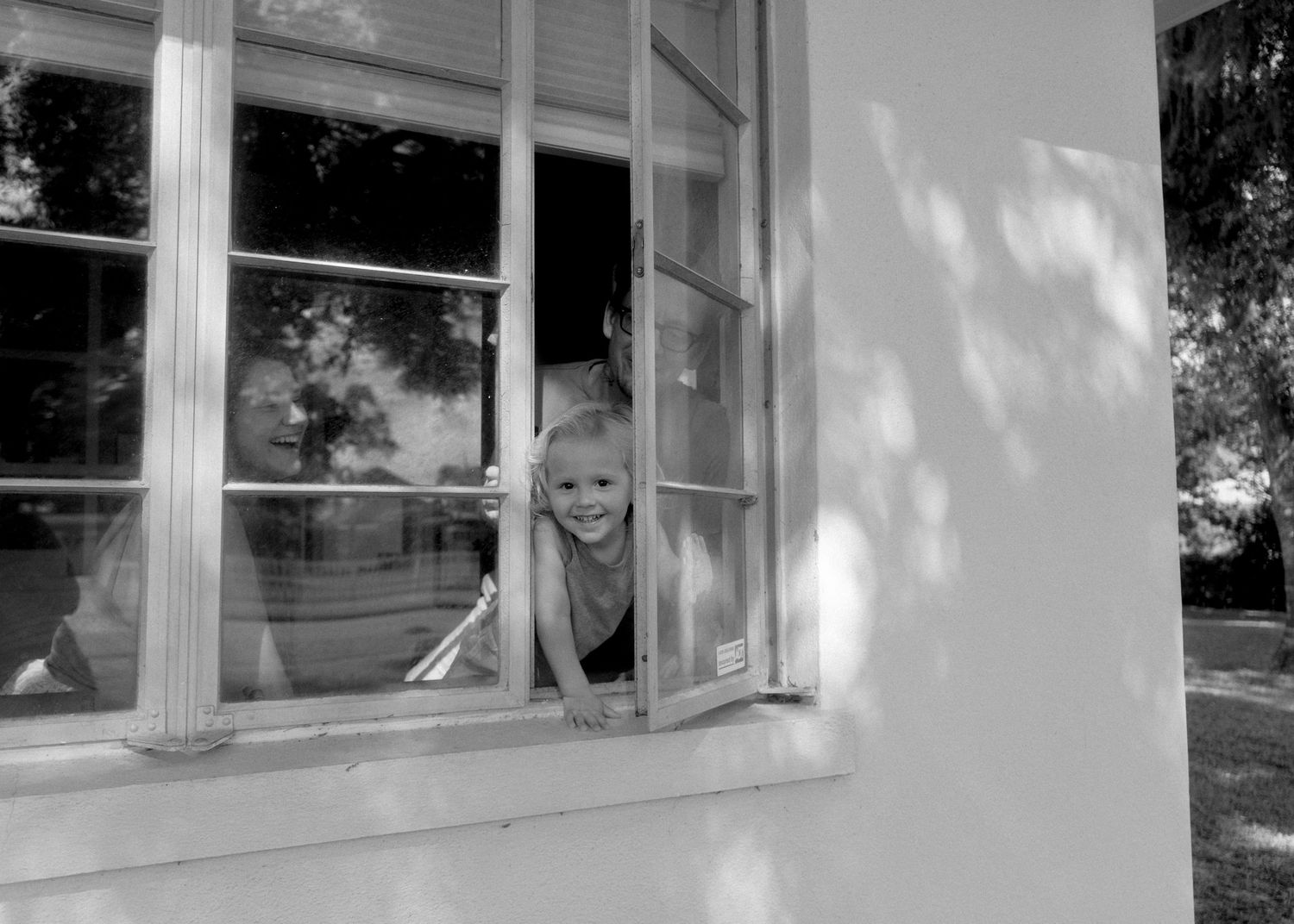 A smiling child peers out of an open window of a white building in this charming black and white photo.