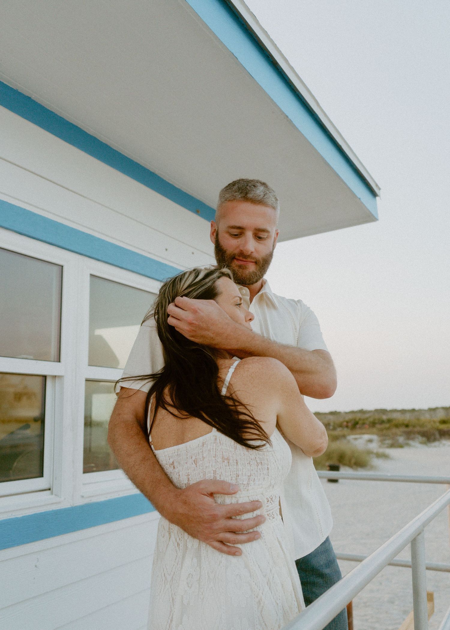 Couple embracing tenderly near a blue and white beach hut at sunset, woman in white dress, man in light shirt.