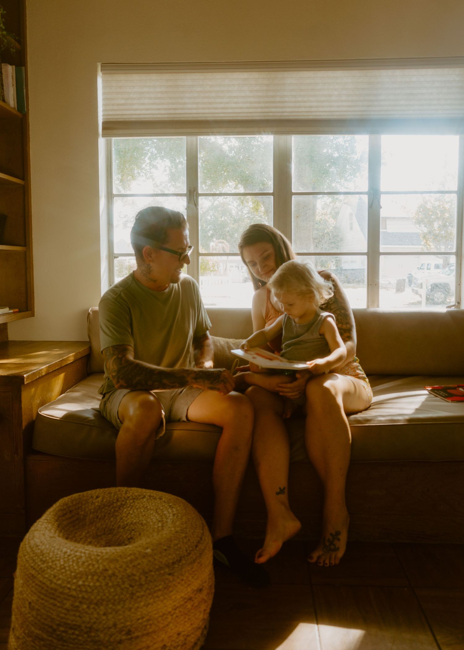 Family of three sitting on a cozy couch reading a book together in warm sunlit living room.