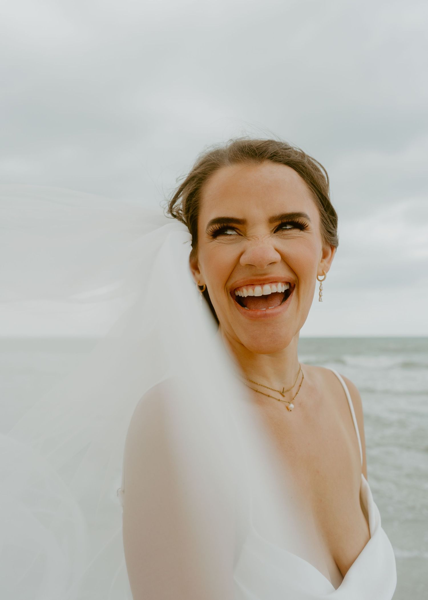 Joyful bride in white gown and veil laughing on a beach with ocean backdrop on her wedding day.