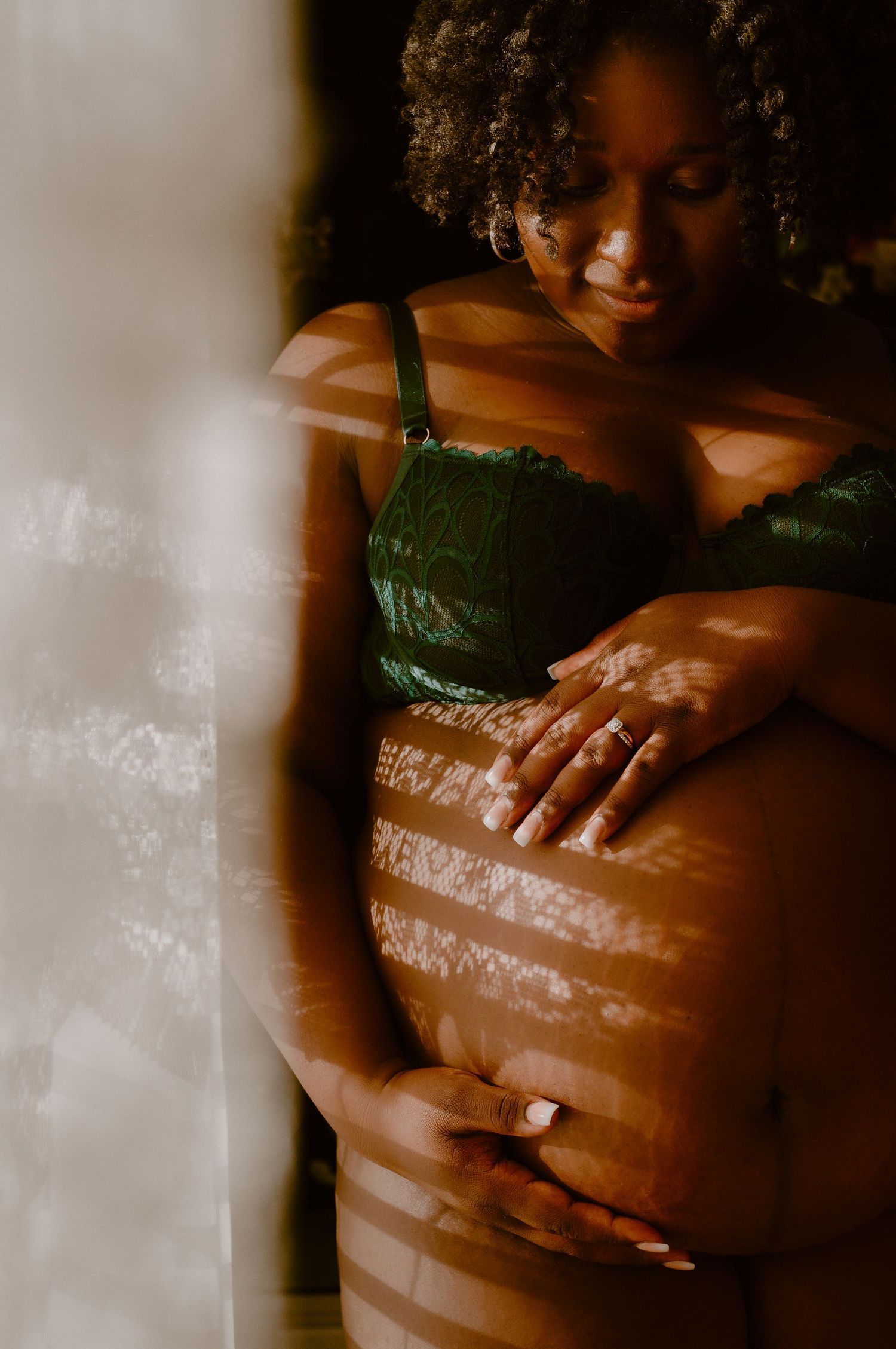 Pregnant Black woman in green lace bralette cradles her baby bump, bathed in warm dappled light.