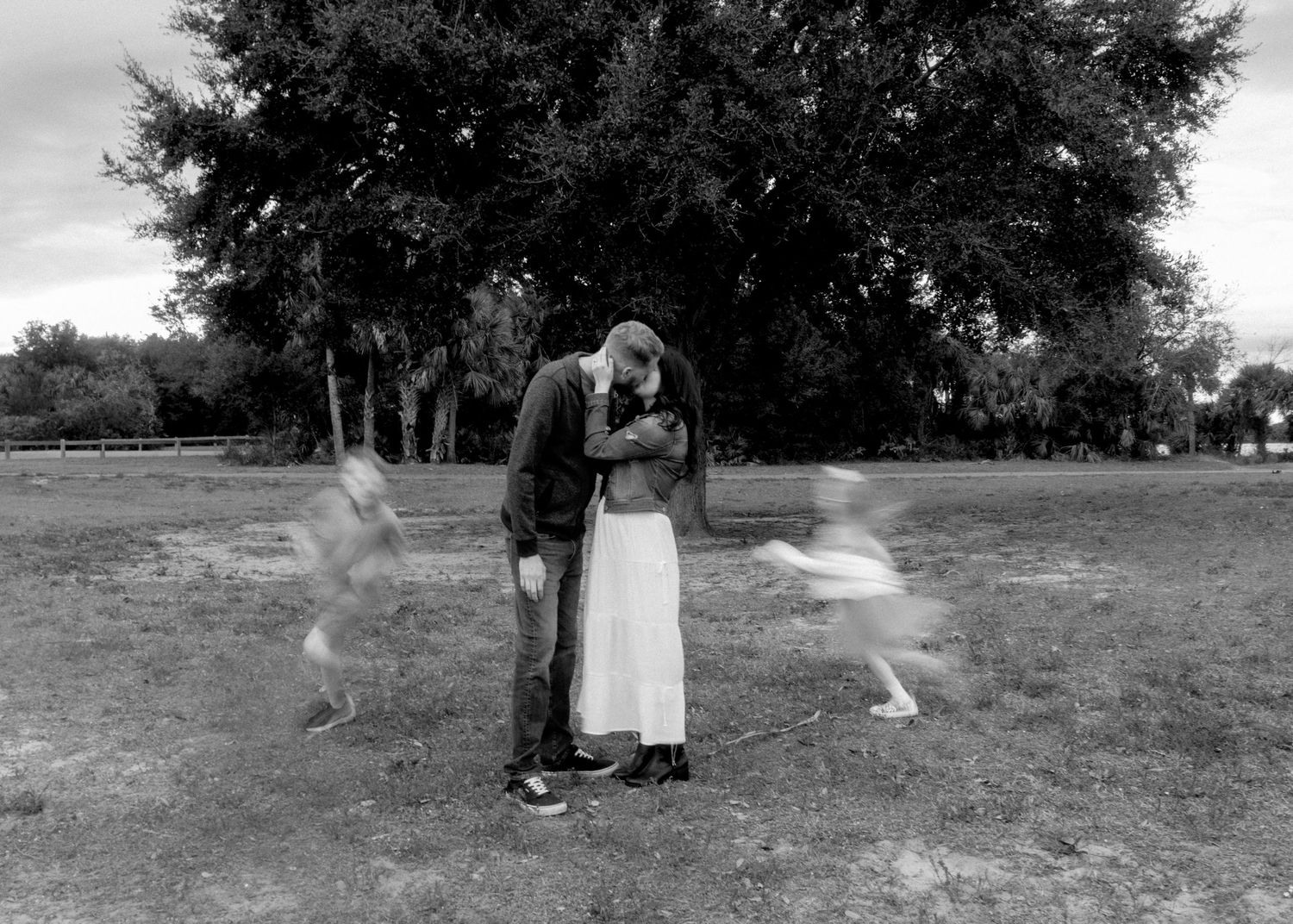 Black and white photo of a couple kissing in a park with blurred children running around them.