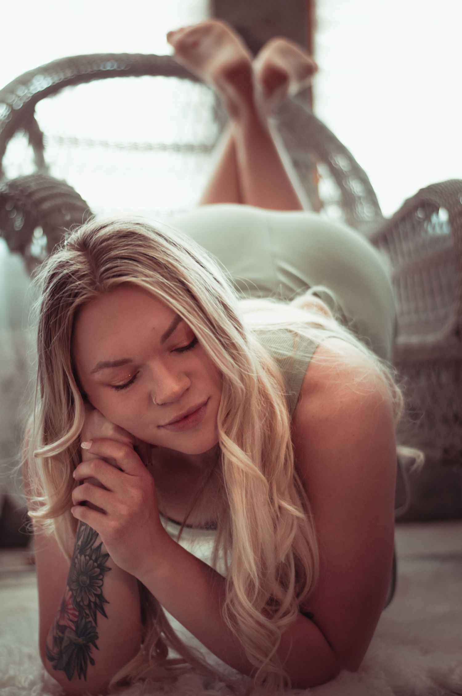 Blonde woman with tattoos lying on floor, eyes closed, legs resting on wicker chair in soft light.