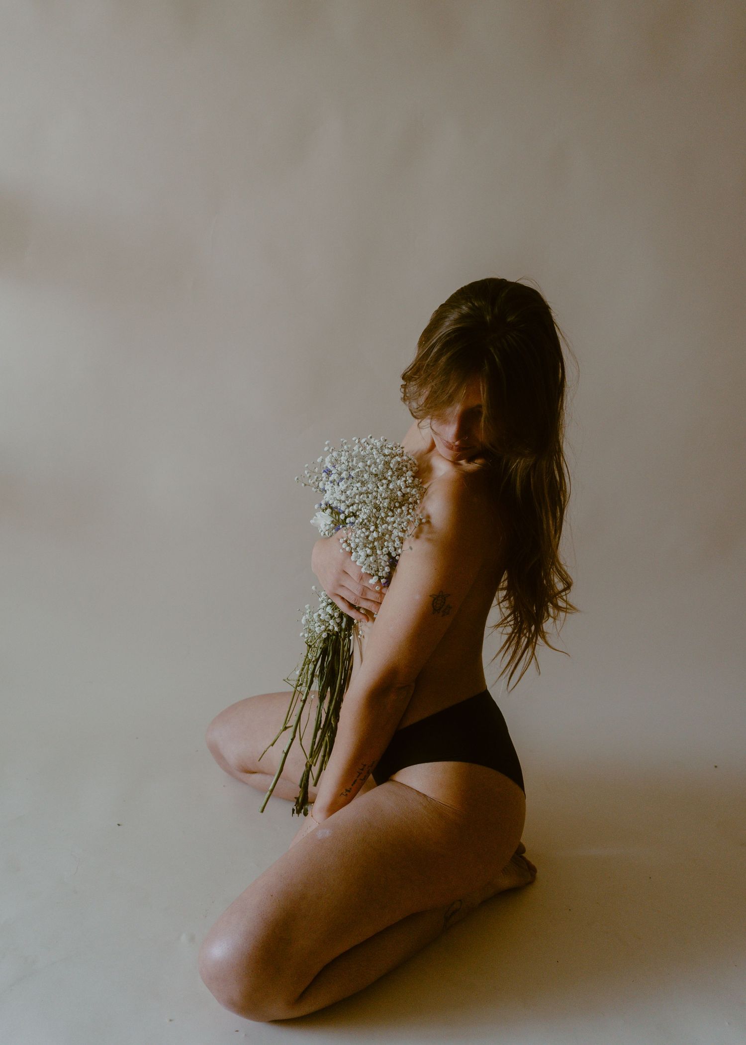 Woman kneeling on floor holding white wildflowers bouquet, wearing black underwear in soft studio lighting.