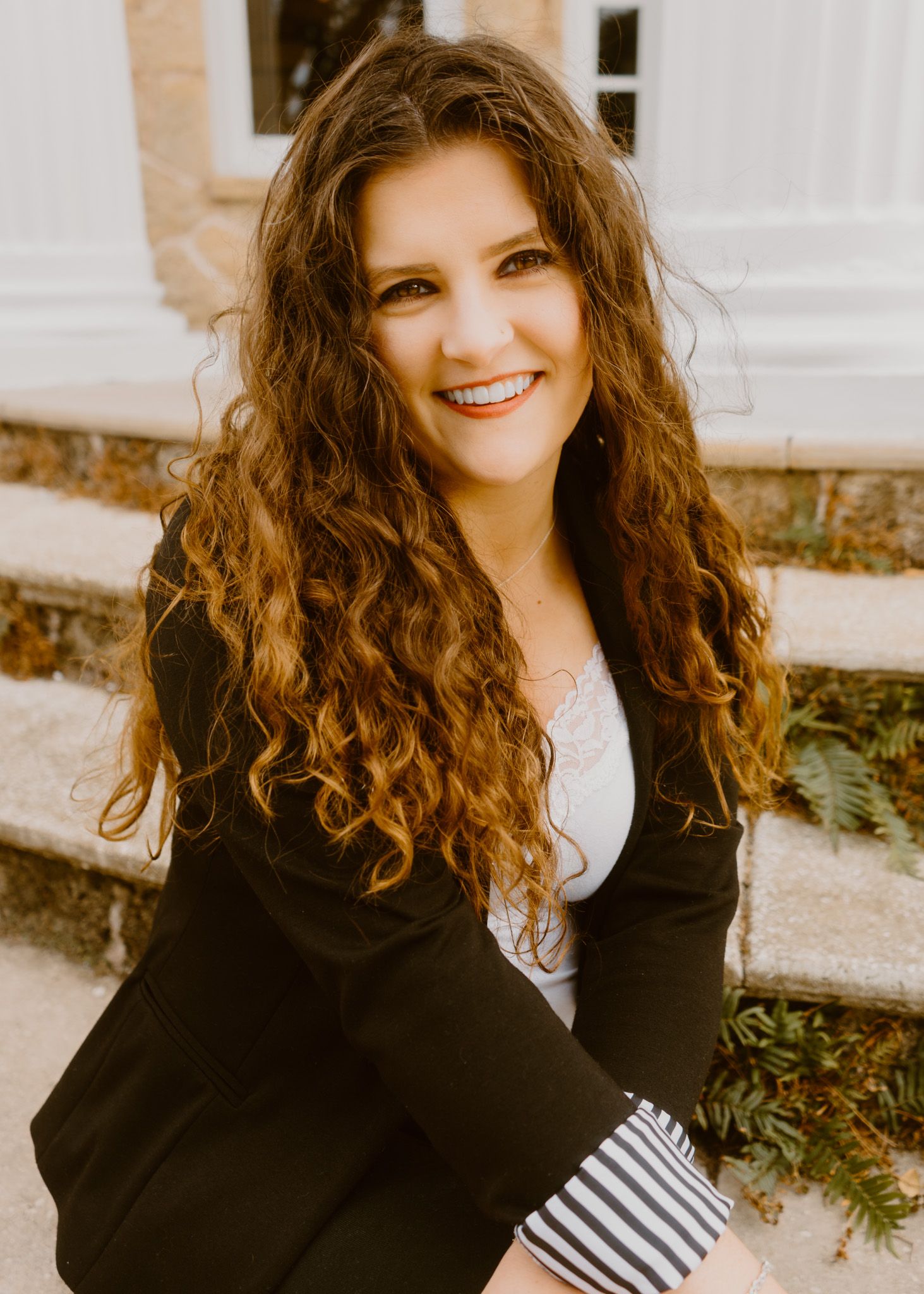 Smiling woman with curly brown hair wearing a black blazer, seated outdoors near stone steps.