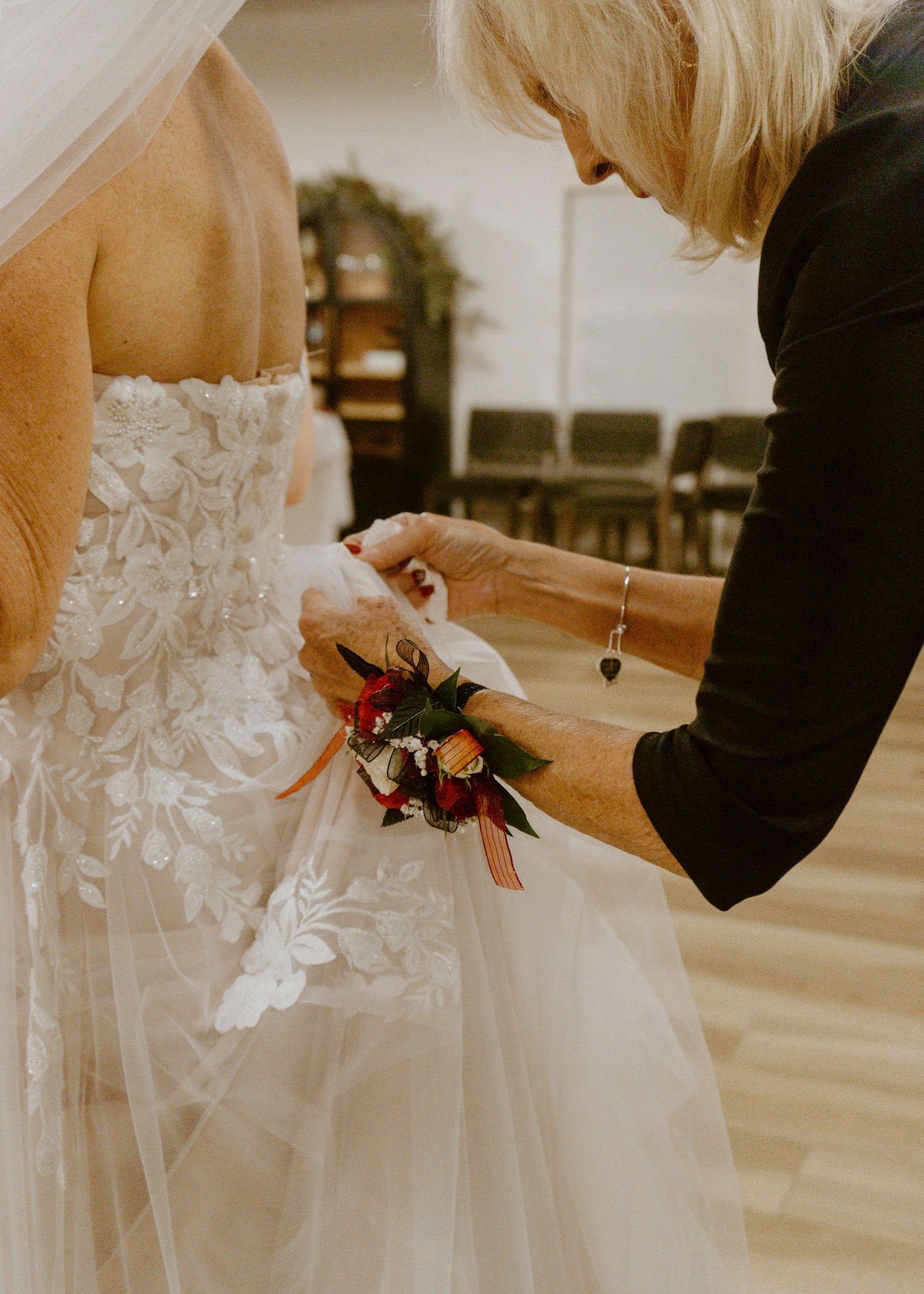Blonde woman helping bride in lace wedding dress with floral wrist corsage before ceremony.
