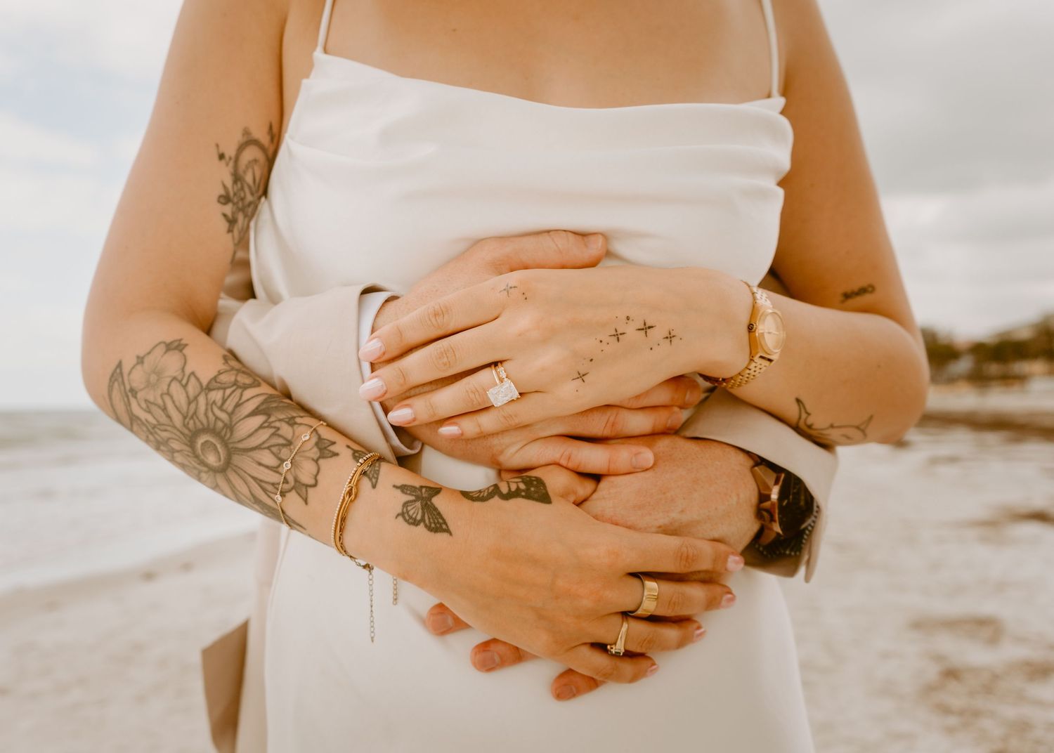 Couple embracing on beach, showing tattooed arms, wedding rings, and gold bracelets, wearing white wedding dress.