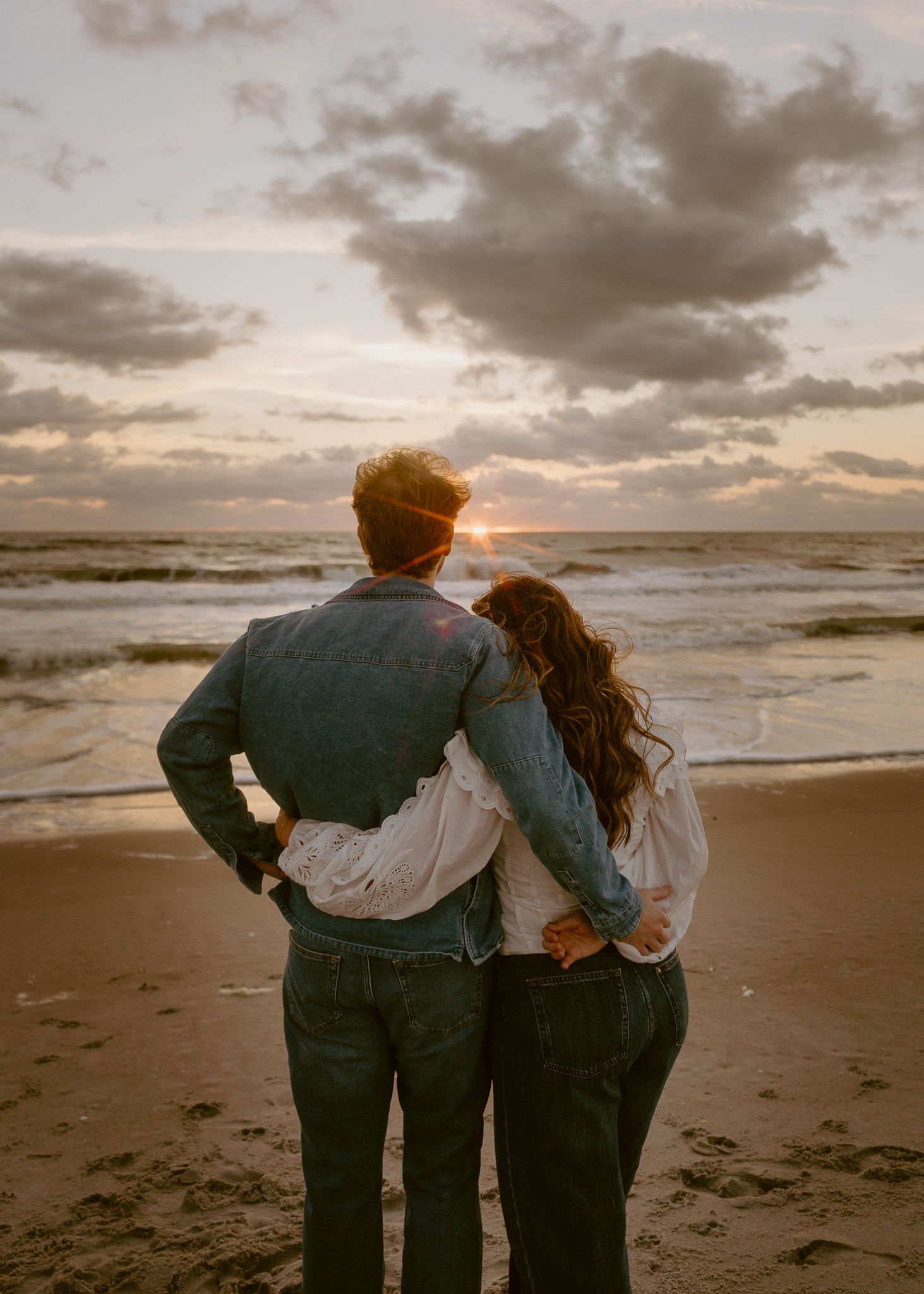 Couple embracing on a sandy beach at sunset, watching ocean waves under a dramatic cloudy sky.