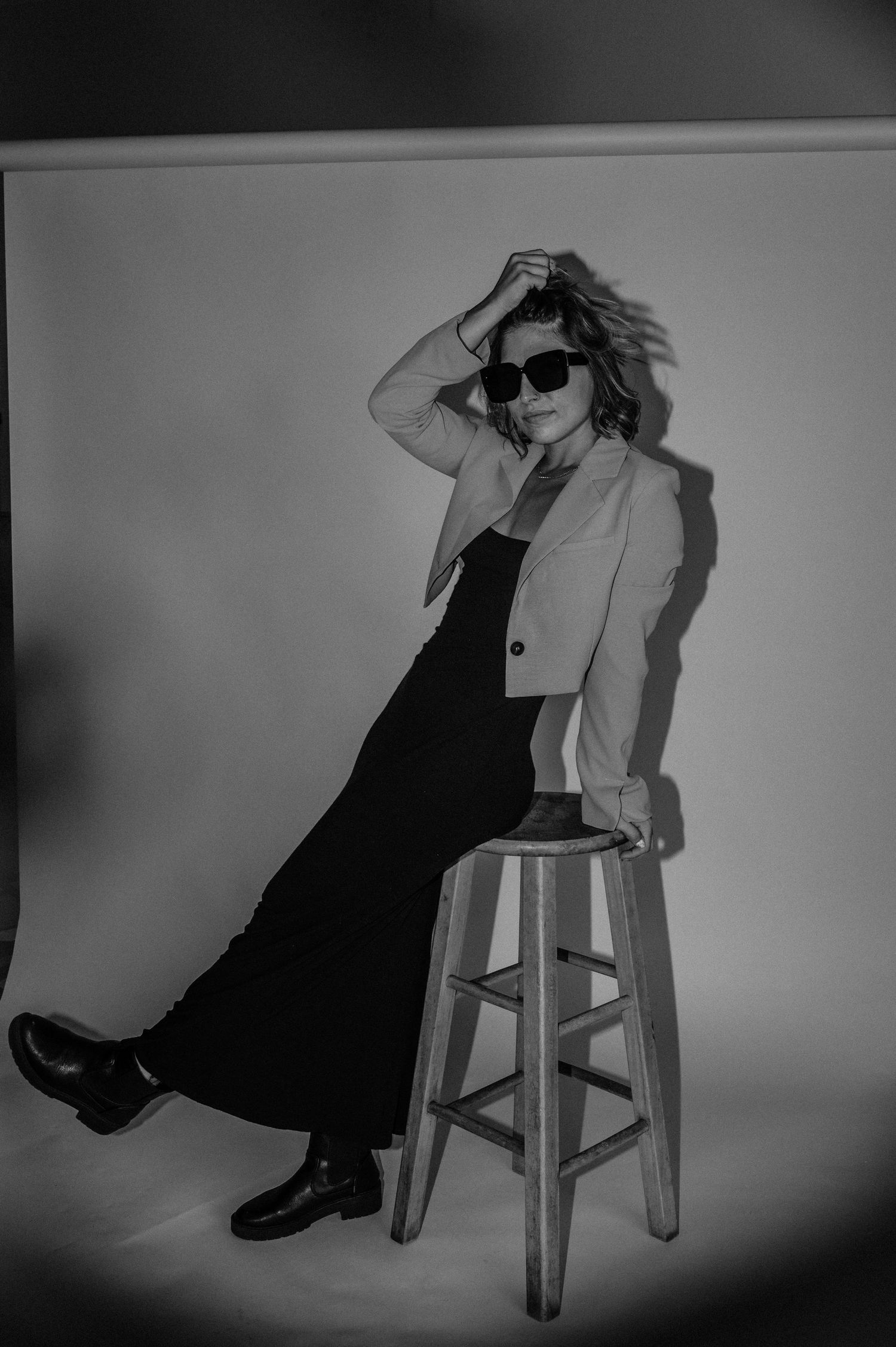 Woman in blazer and sunglasses posing on wooden stool in black and white studio fashion photo.