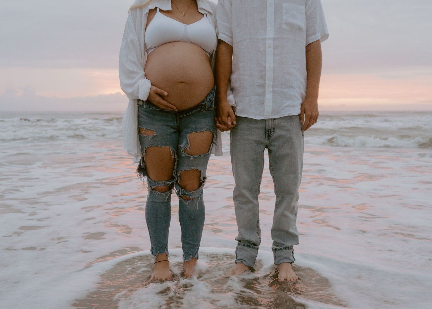 Couple standing barefoot in ocean waves, pregnant woman showing baby bump in ripped jeans at sunset.
