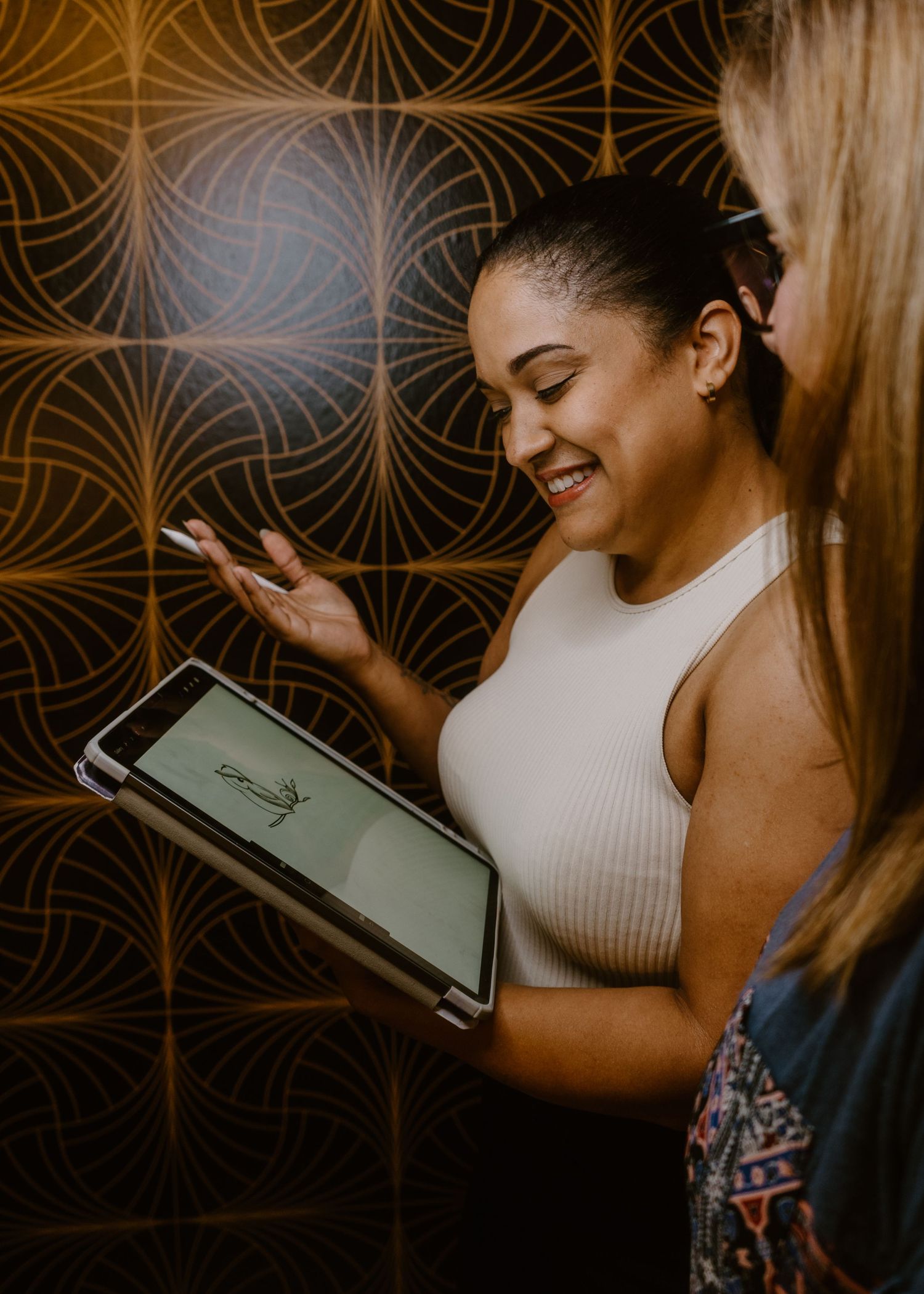 Two women smiling and sharing content on a tablet device against a decorative patterned wall.